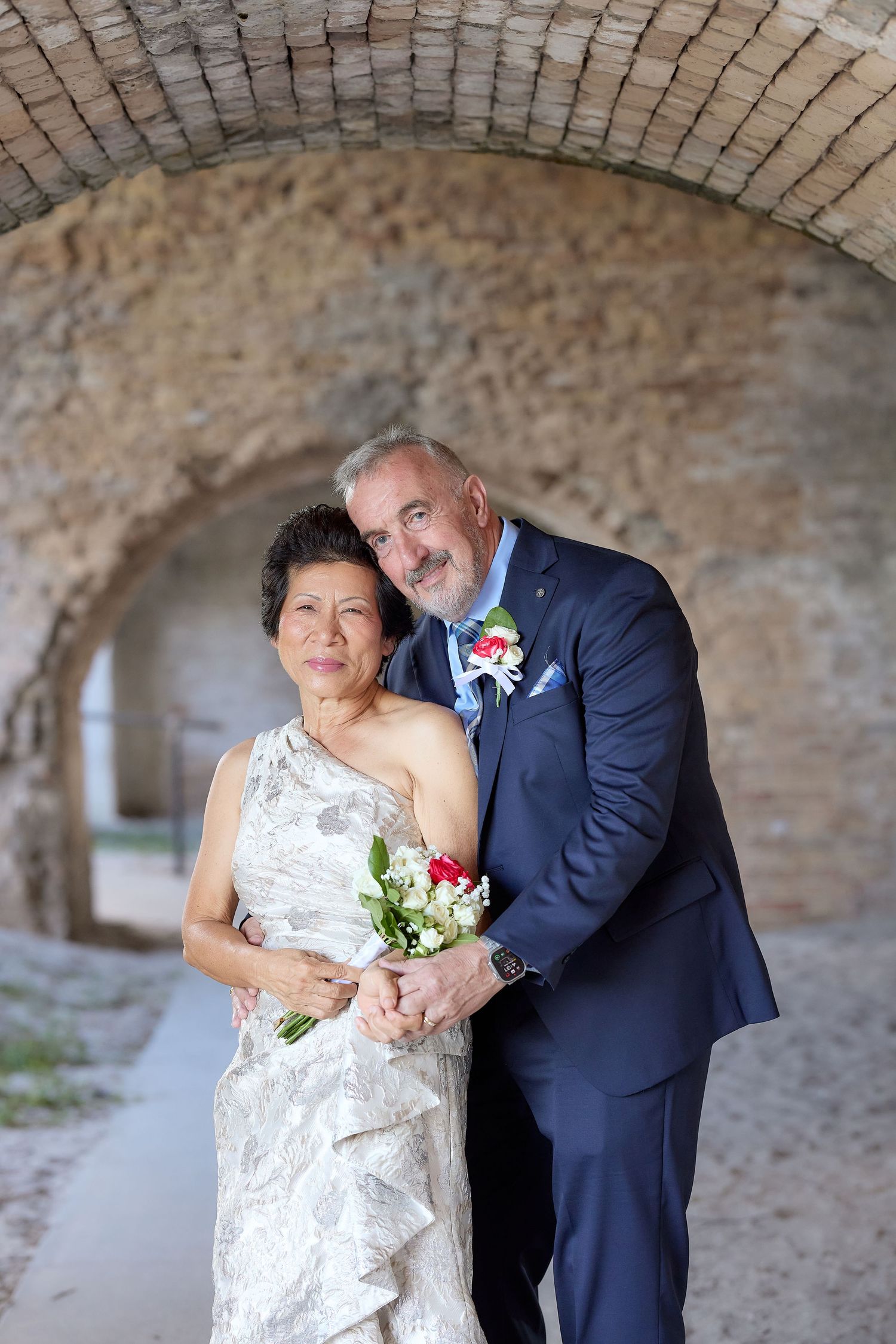 Mature interracial couple in wedding attire posing under stone arch, bride holding bouquet, groom in navy suit.