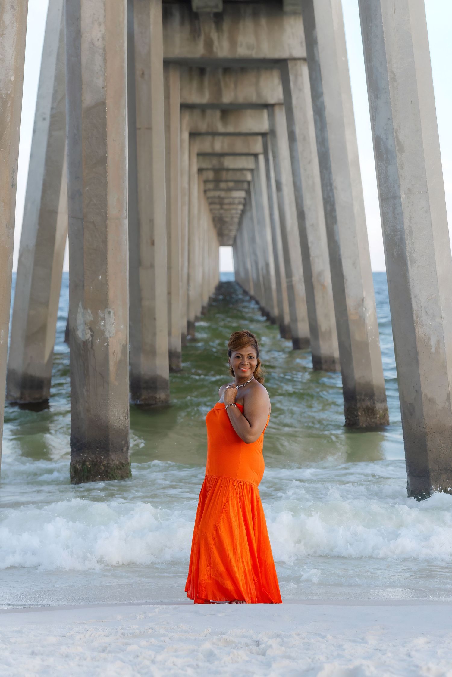 Woman in vibrant orange dress stands under a long beach pier with ocean waves surrounding her.