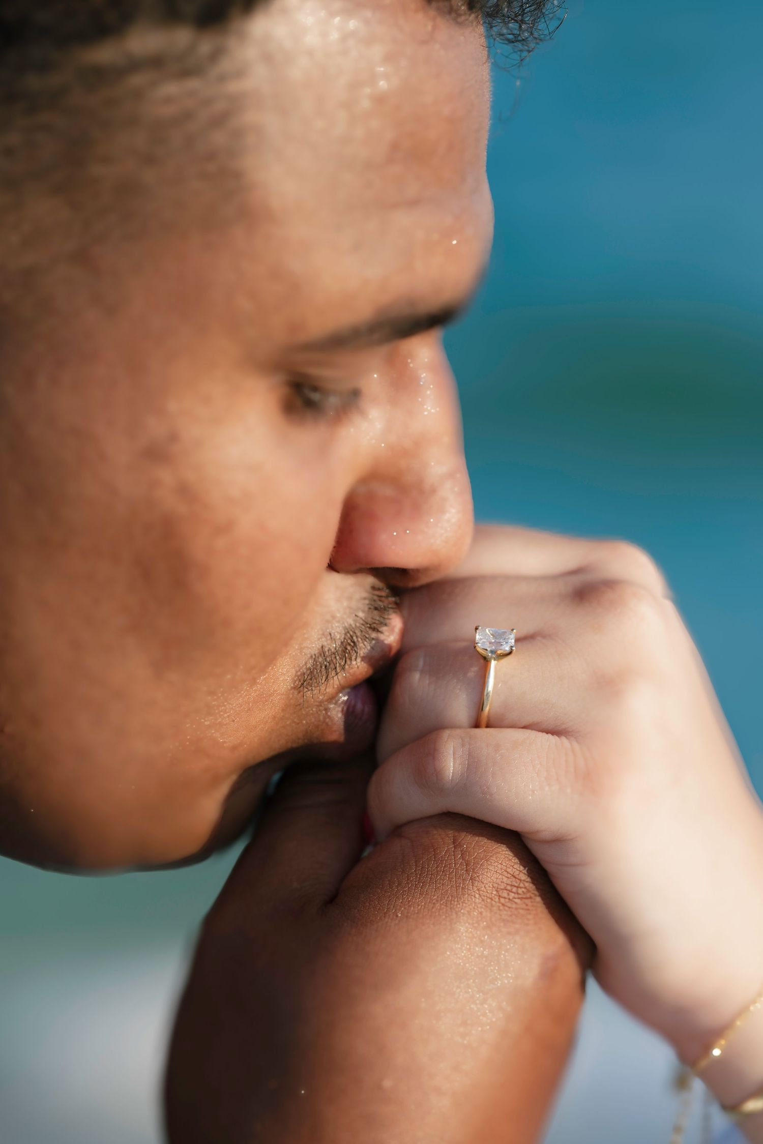 Man kissing hand of woman wearing diamond engagement ring near ocean beach proposal.