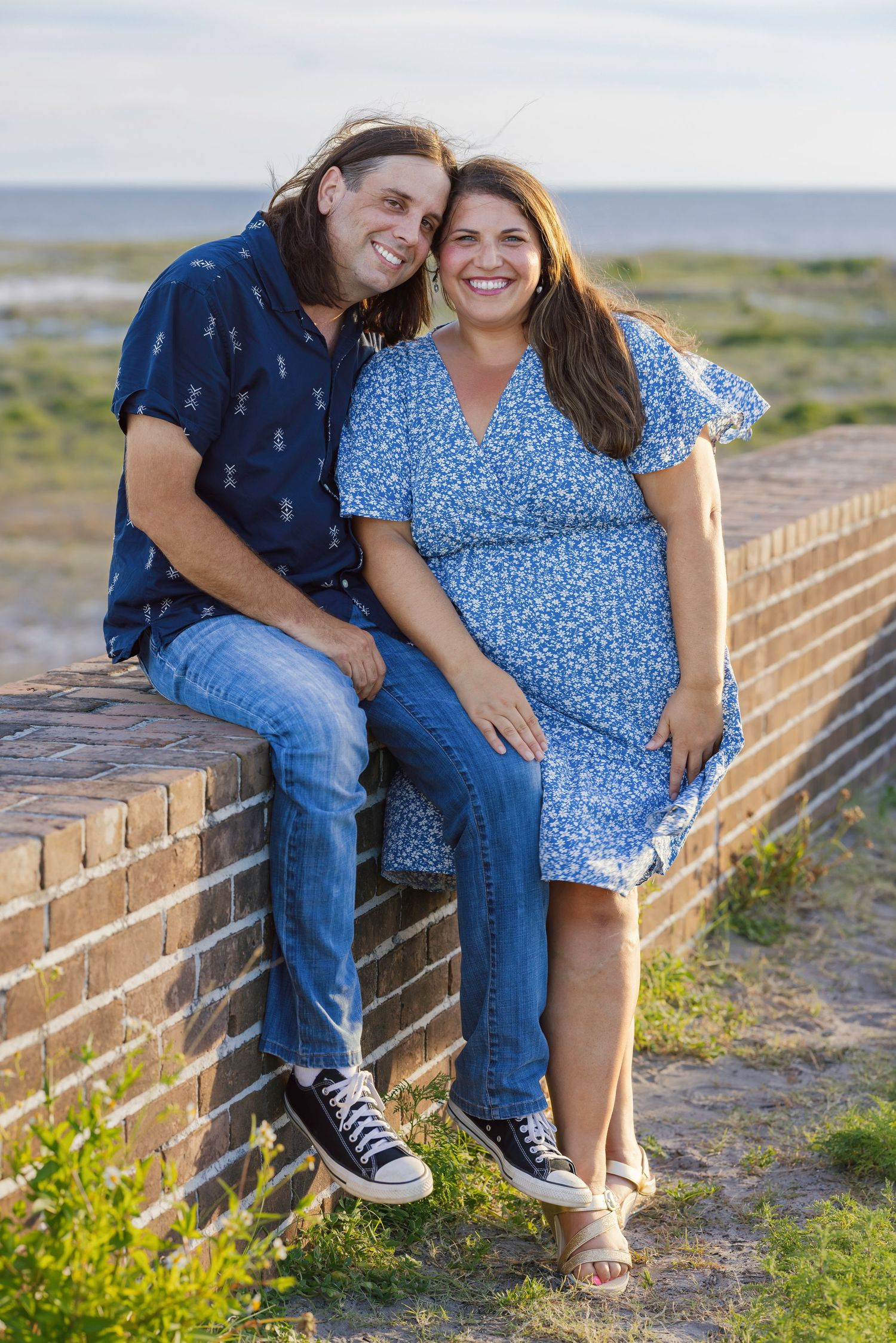Smiling couple sitting on a brick wall near the beach, wearing blue outfits, enjoying a sunny day outdoors.