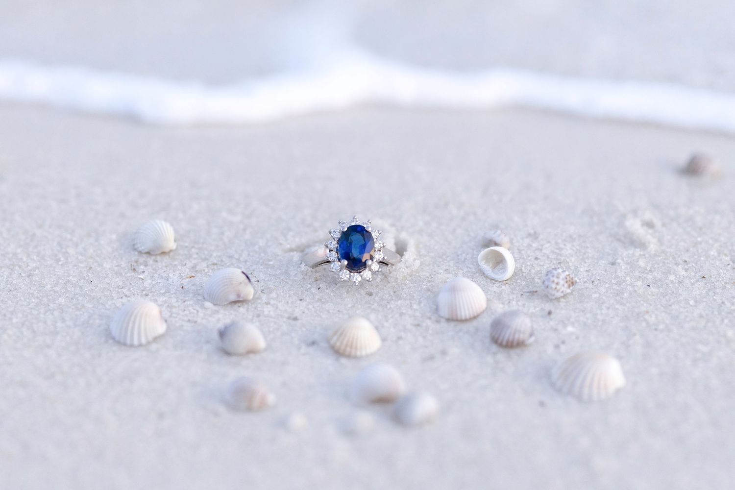 Blue sapphire halo engagement ring resting on white sand beach surrounded by small seashells with ocean waves in background.