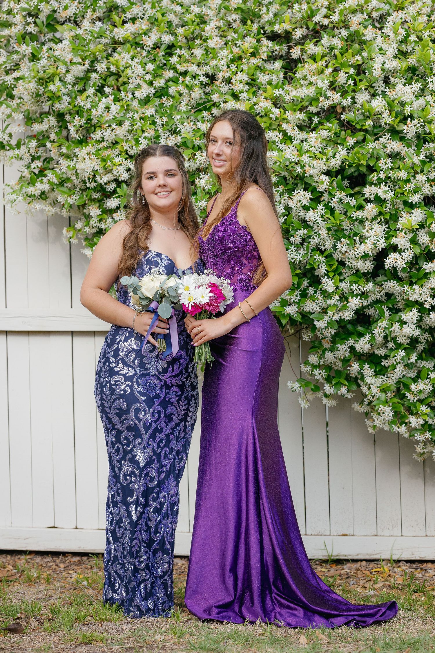 Two ladies in navy and purple formal dresses pose together against a white fence with blooming flowers.