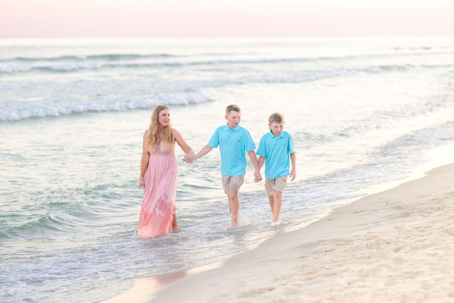 People wearing pink and aqua blue clothing walk hand in hand along a beach at sunset with ocean waves rolling in.