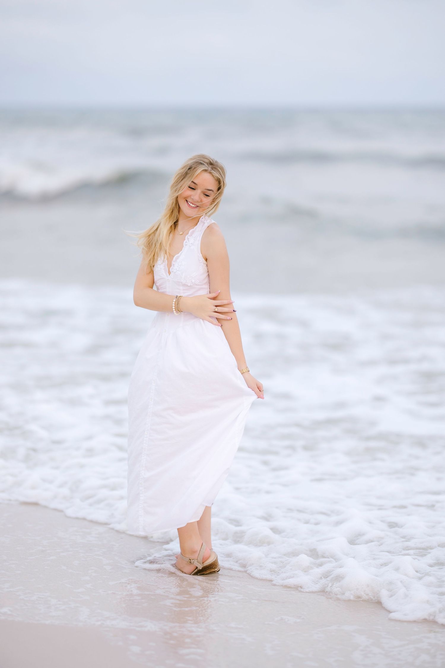 Woman in flowing white dress standing at the edge of ocean waves on a misty beach.