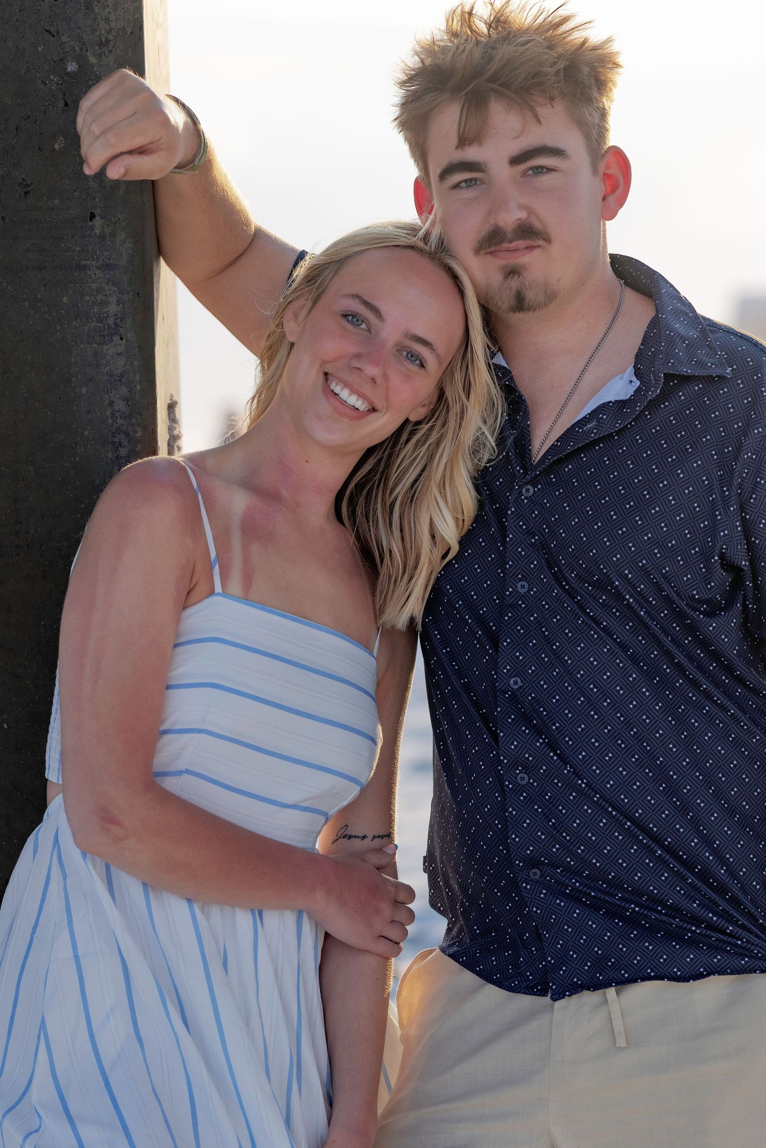 Happy couple leaning against a pier post at sunset, woman in striped sundress and man in navy shirt.