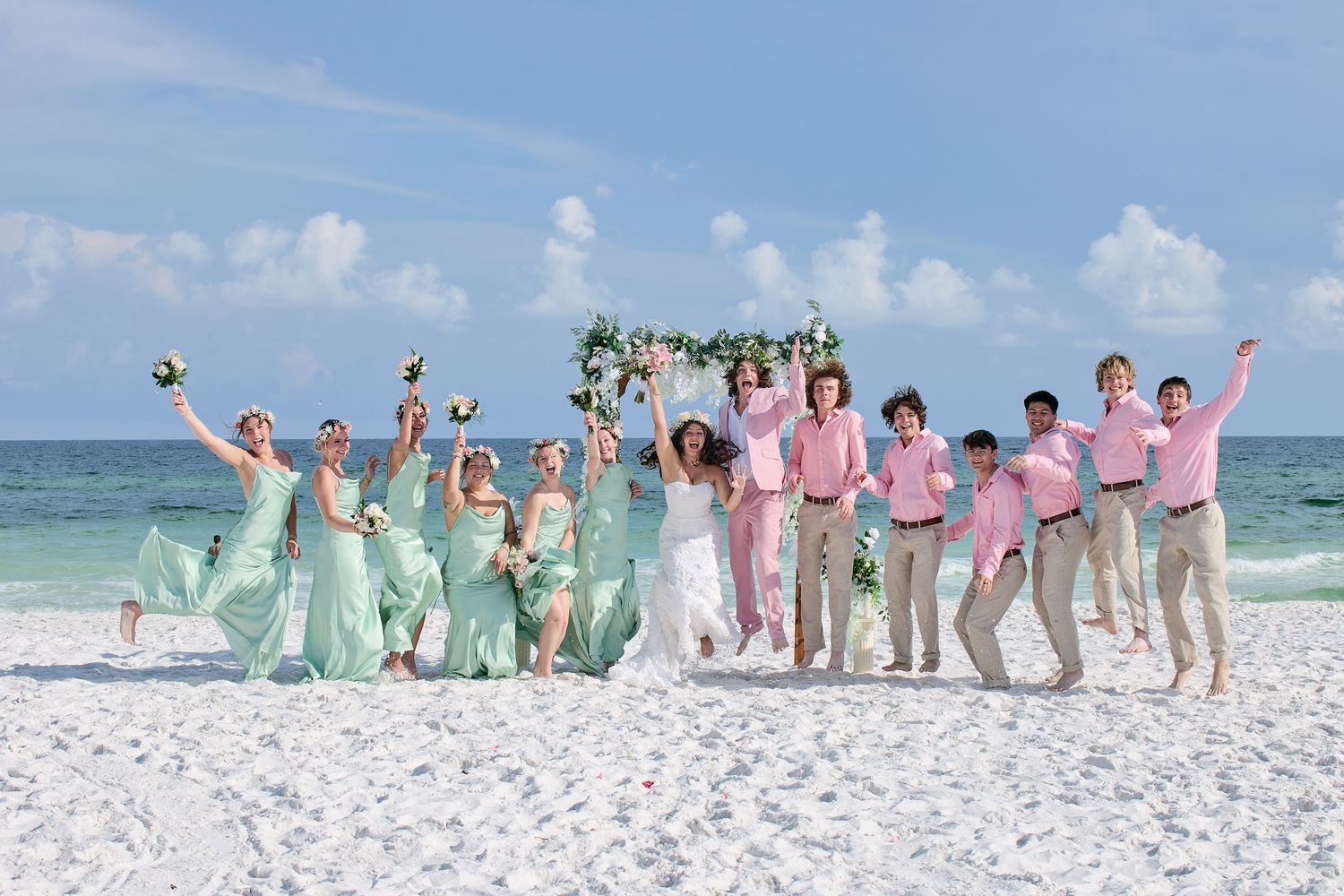 Joyful wedding party jumping on white sand beach, bridesmaids in mint green and groomsmen in pink.