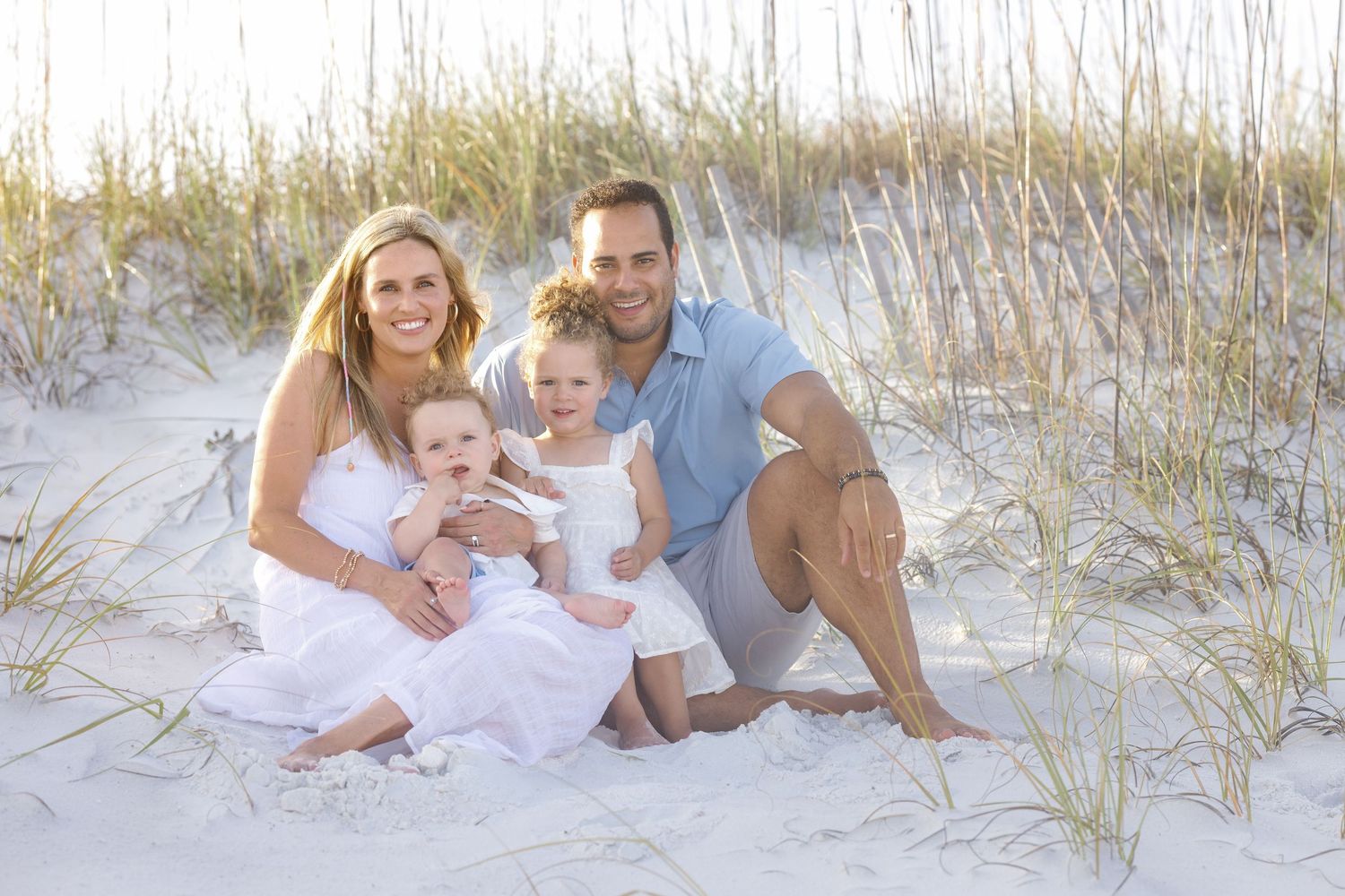 Happy family of four sitting together on white sand beach dunes, parents with two young children in white outfits.