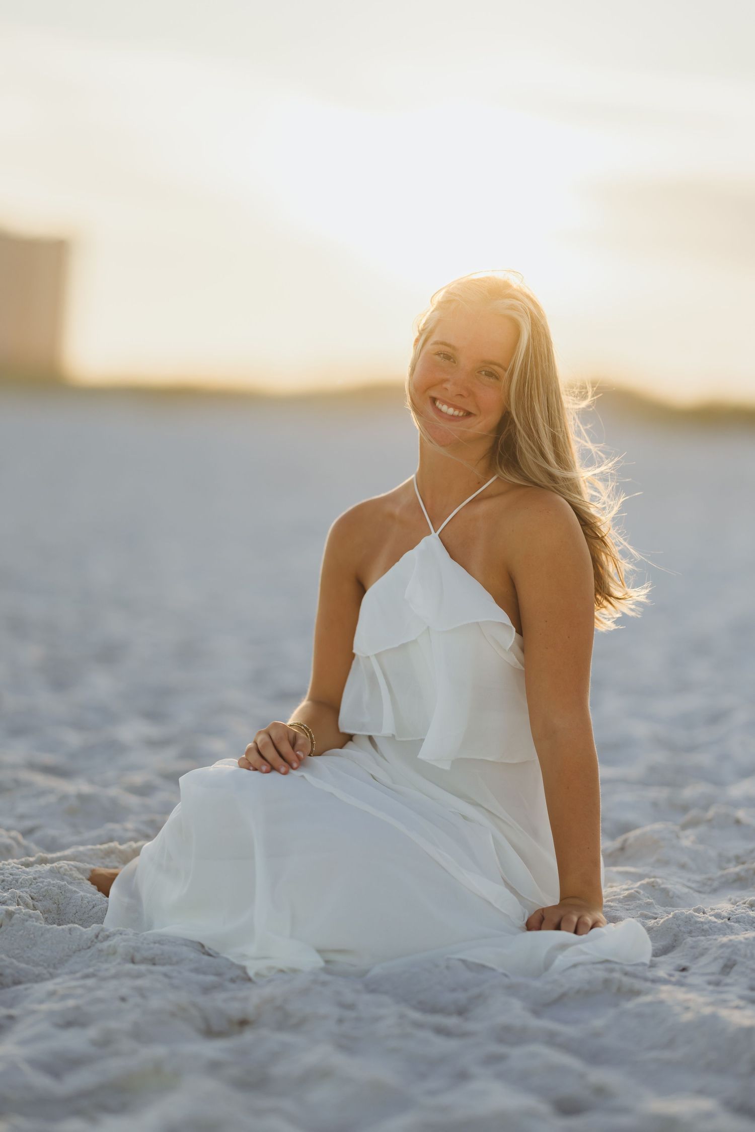 Smiling blonde woman in white halter dress sitting on sandy beach at golden sunset.