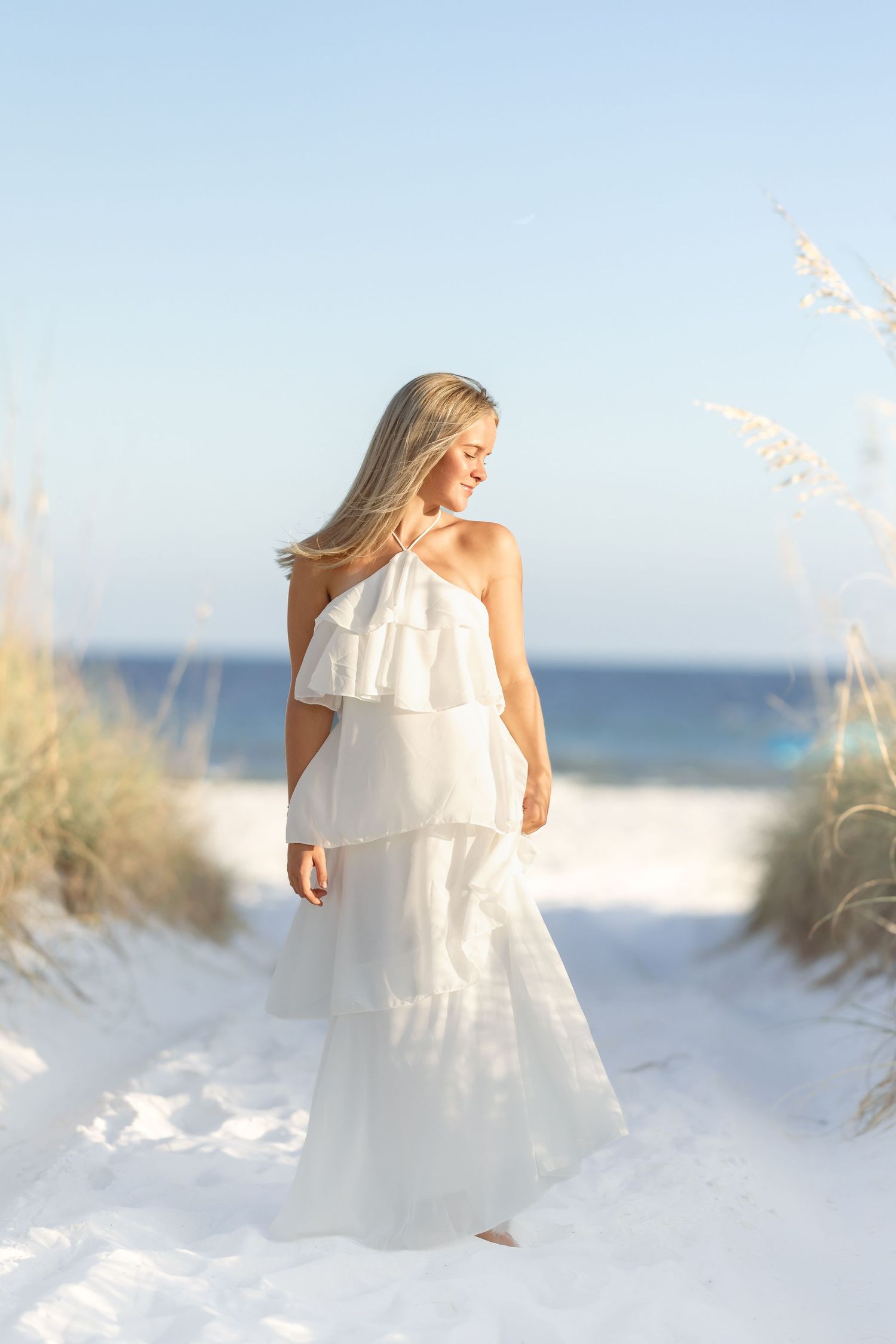 Blonde woman in white tiered halter maxi dress standing on sandy beach path with ocean in background.