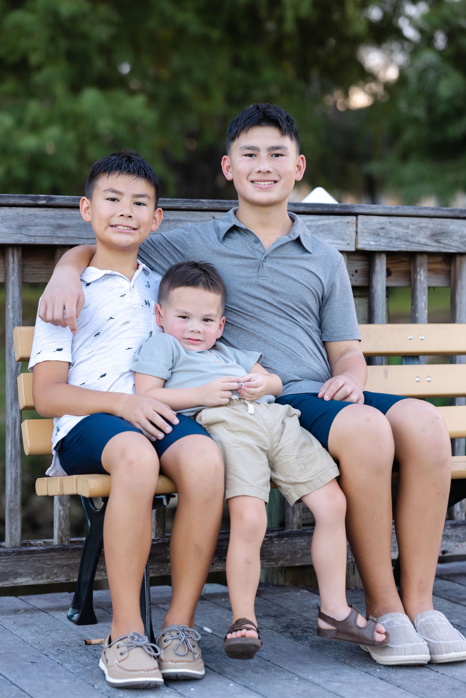 Three young boys smiling and posing together on a wooden bench outdoors in a park setting.