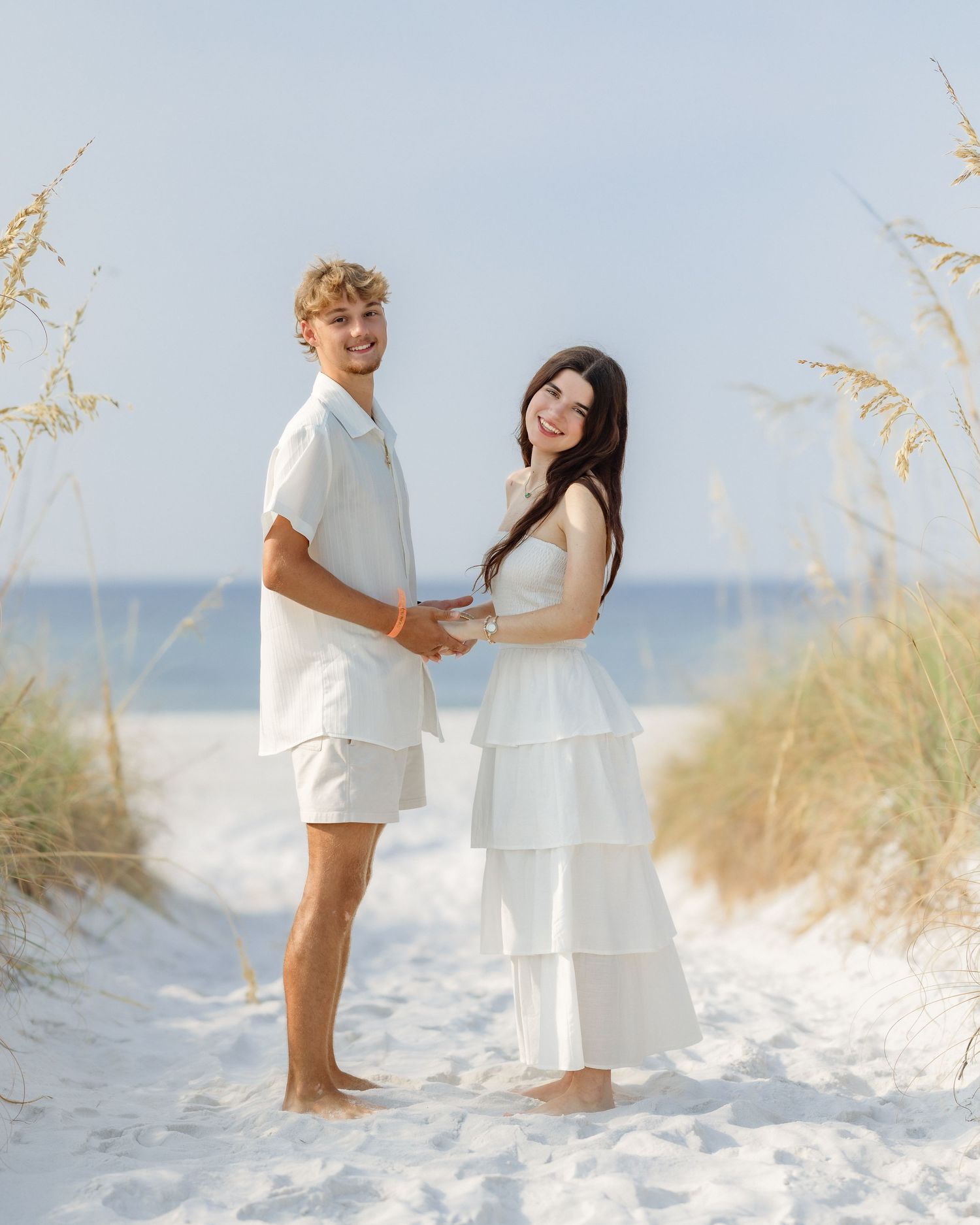 Young couple in matching white outfits holding hands on a sandy beach path surrounded by sea oats.