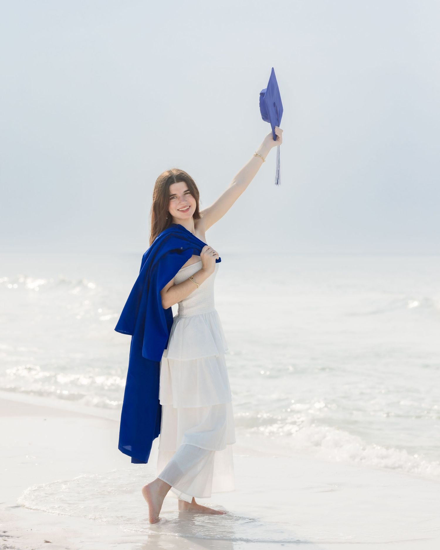 Woman in white dress holding blue graduation cap and gown on a bright beach shoreline.