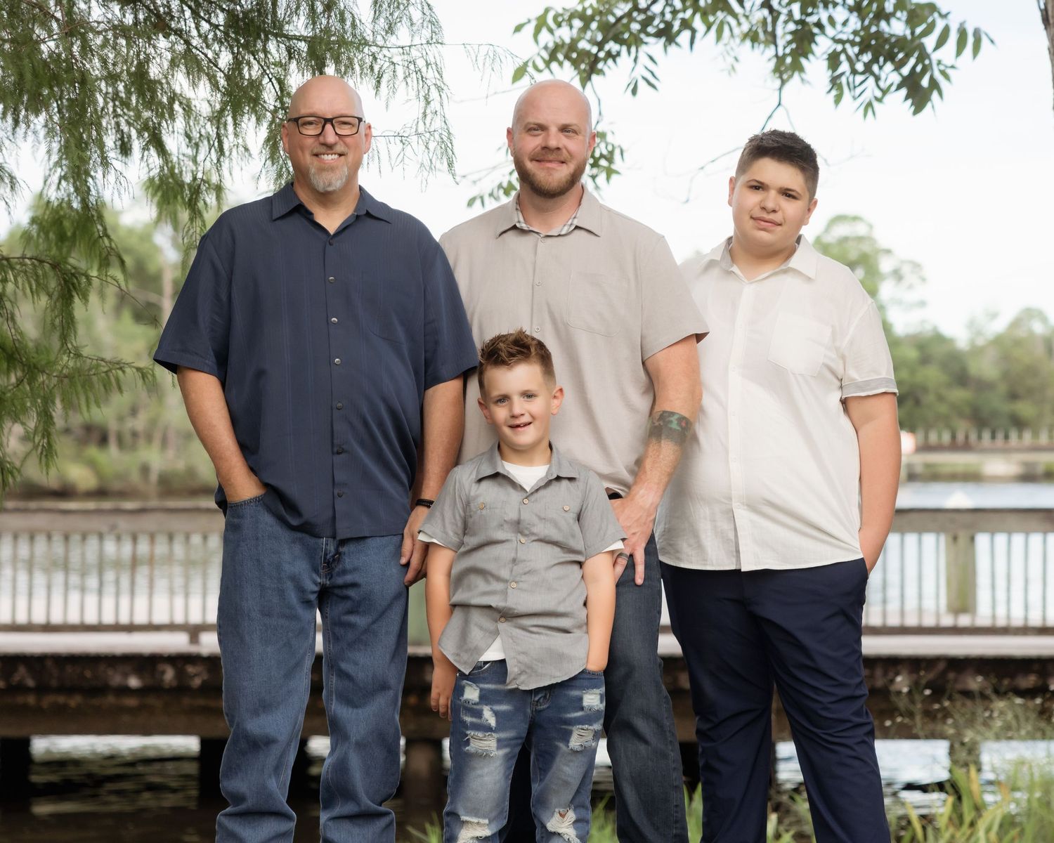 Family of four posing outdoors near a lake with willow trees in a warm, natural setting.