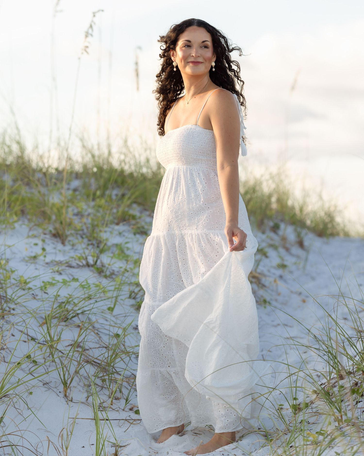 Woman in white strapless maxi dress standing on sandy beach dunes with sea oats in background.