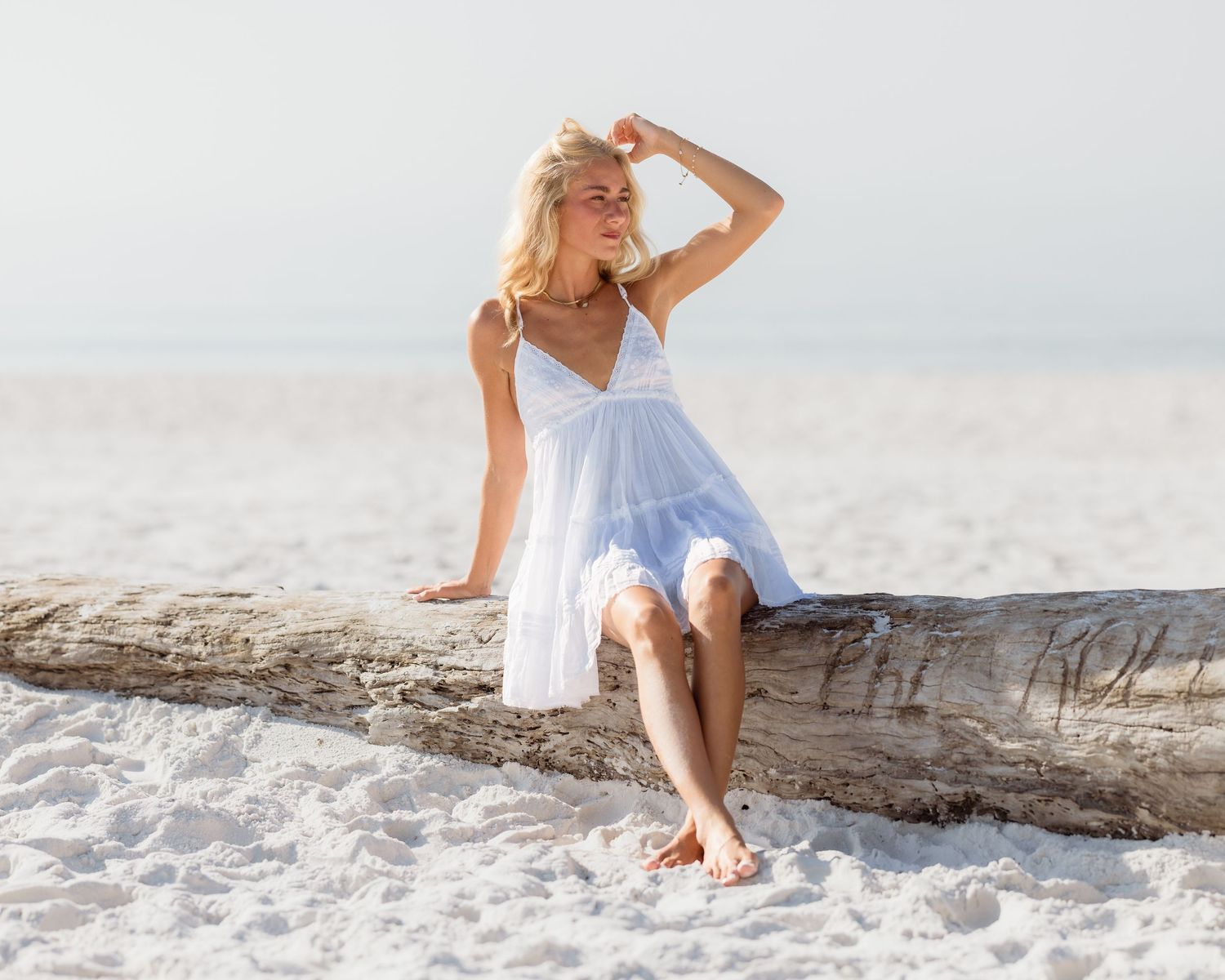 Blonde woman in white sundress sitting on driftwood log at sandy beach on sunny day.