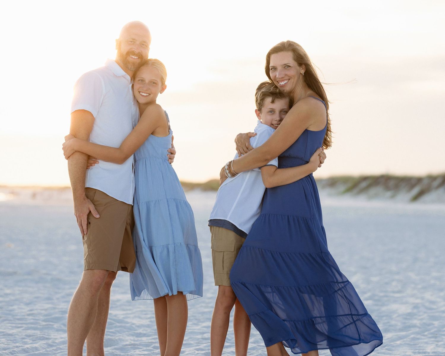 Family of four posing on a white sand beach at sunset, wearing coordinating blue and white outfits.