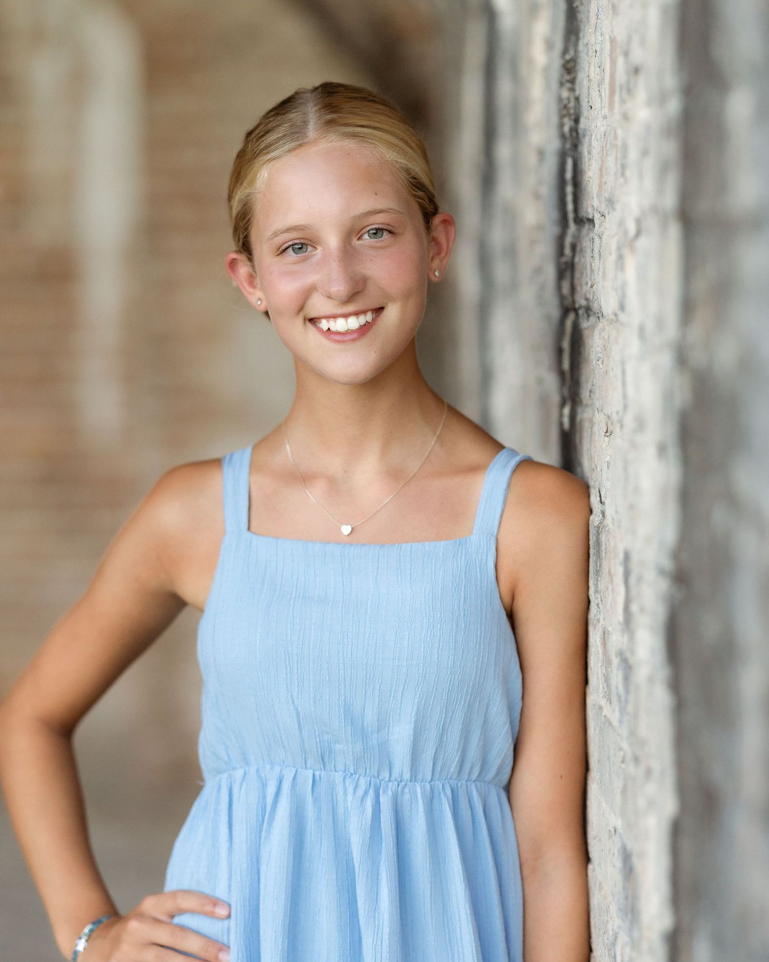 Smiling teenage girl in a light blue sundress leaning against a brick wall outdoors.