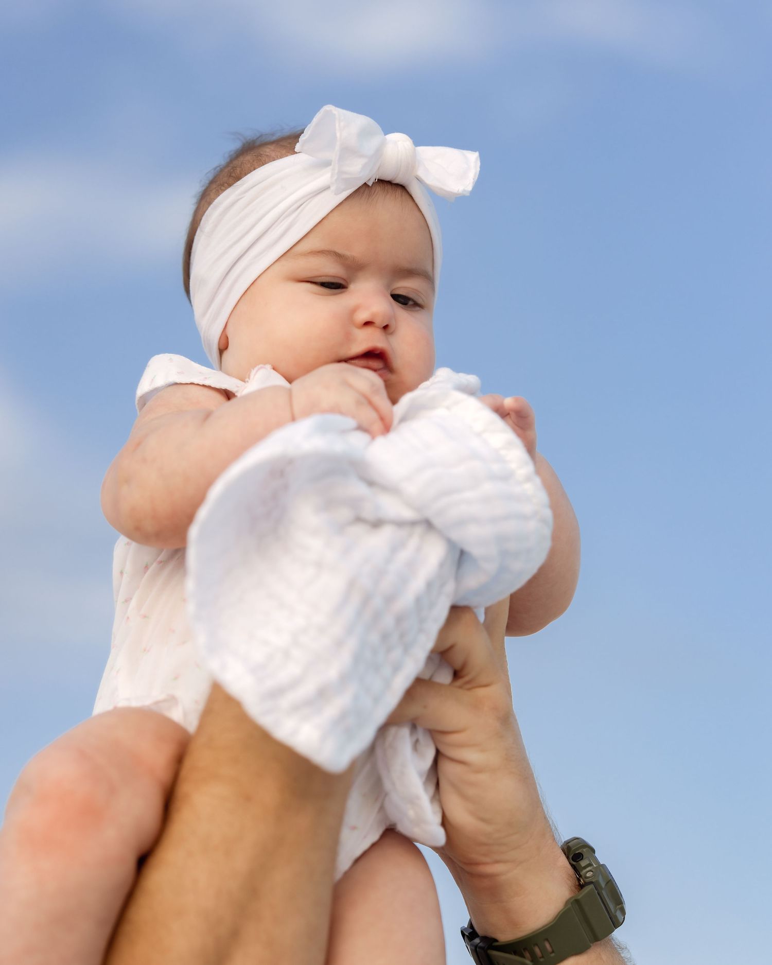 Adorable baby in white outfit and bow headband lifted against a blue sky outdoors.