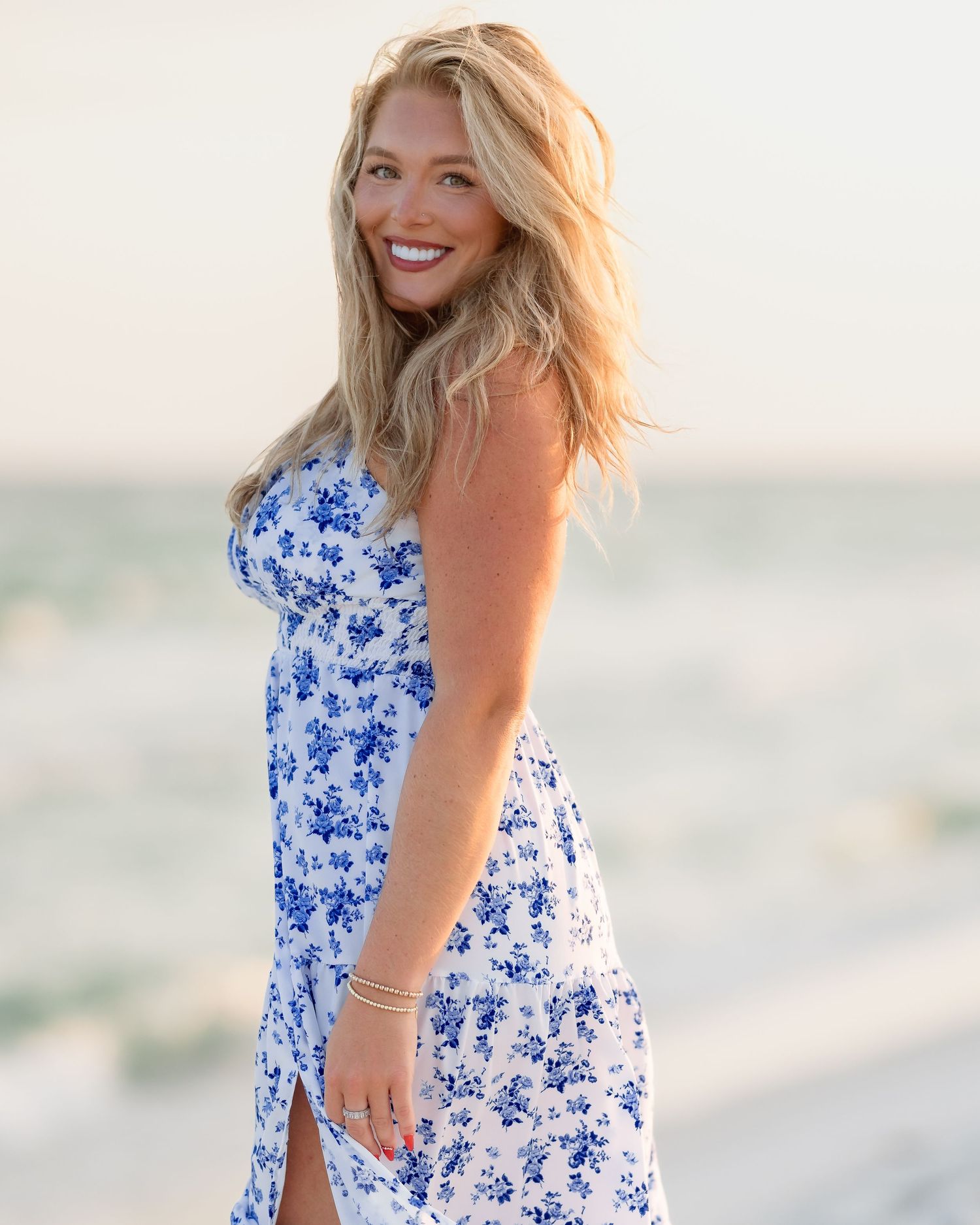 Smiling blonde woman in blue floral sundress posing on sunny beach near ocean.