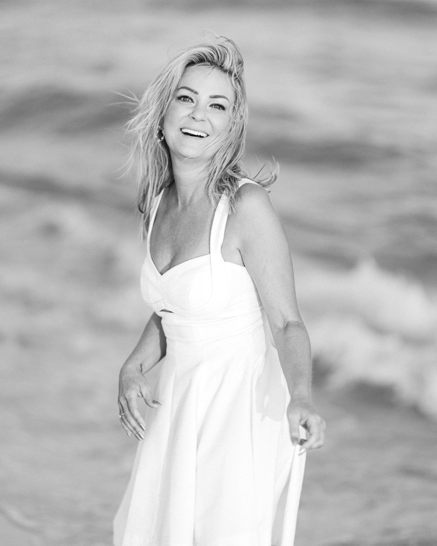 Black and white photo of a laughing blonde woman in white dress standing by ocean waves.