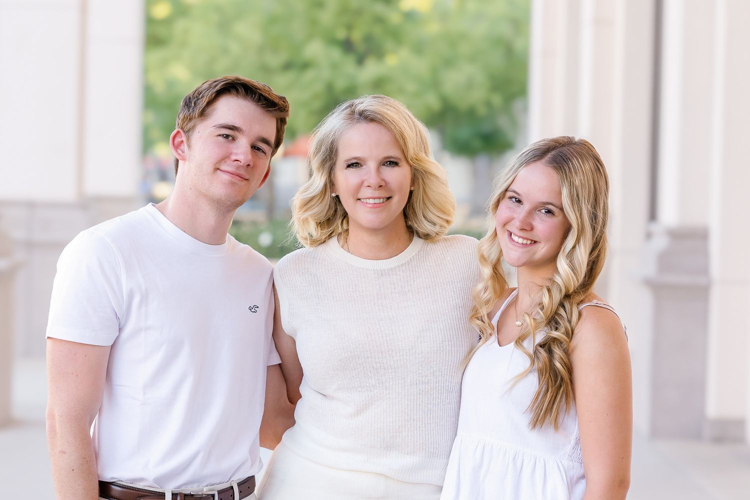 Three smiling young adults in white outfits posing together outdoors near elegant columns.