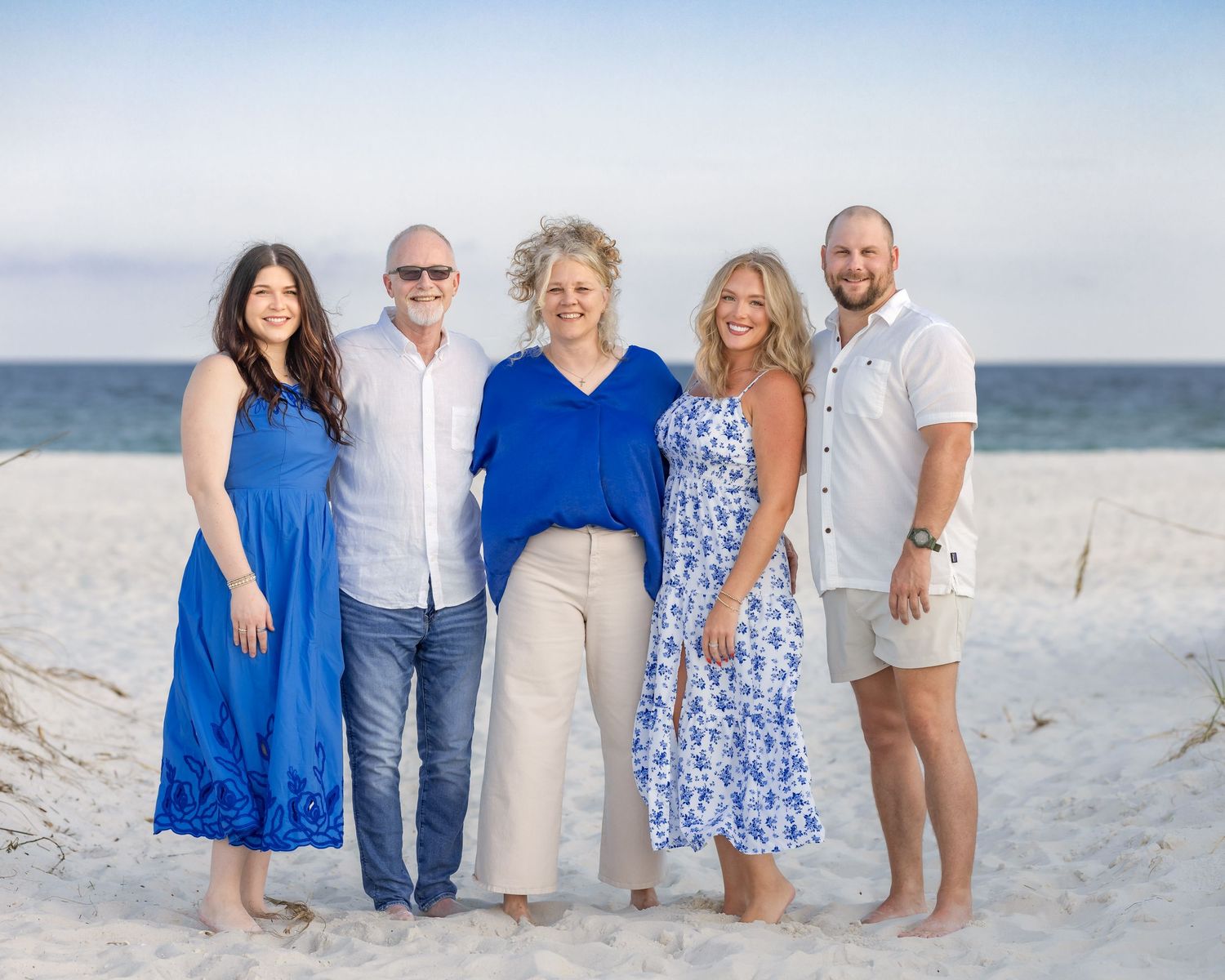 Family of five smiling together on a sandy beach with ocean backdrop, wearing blue and white outfits.