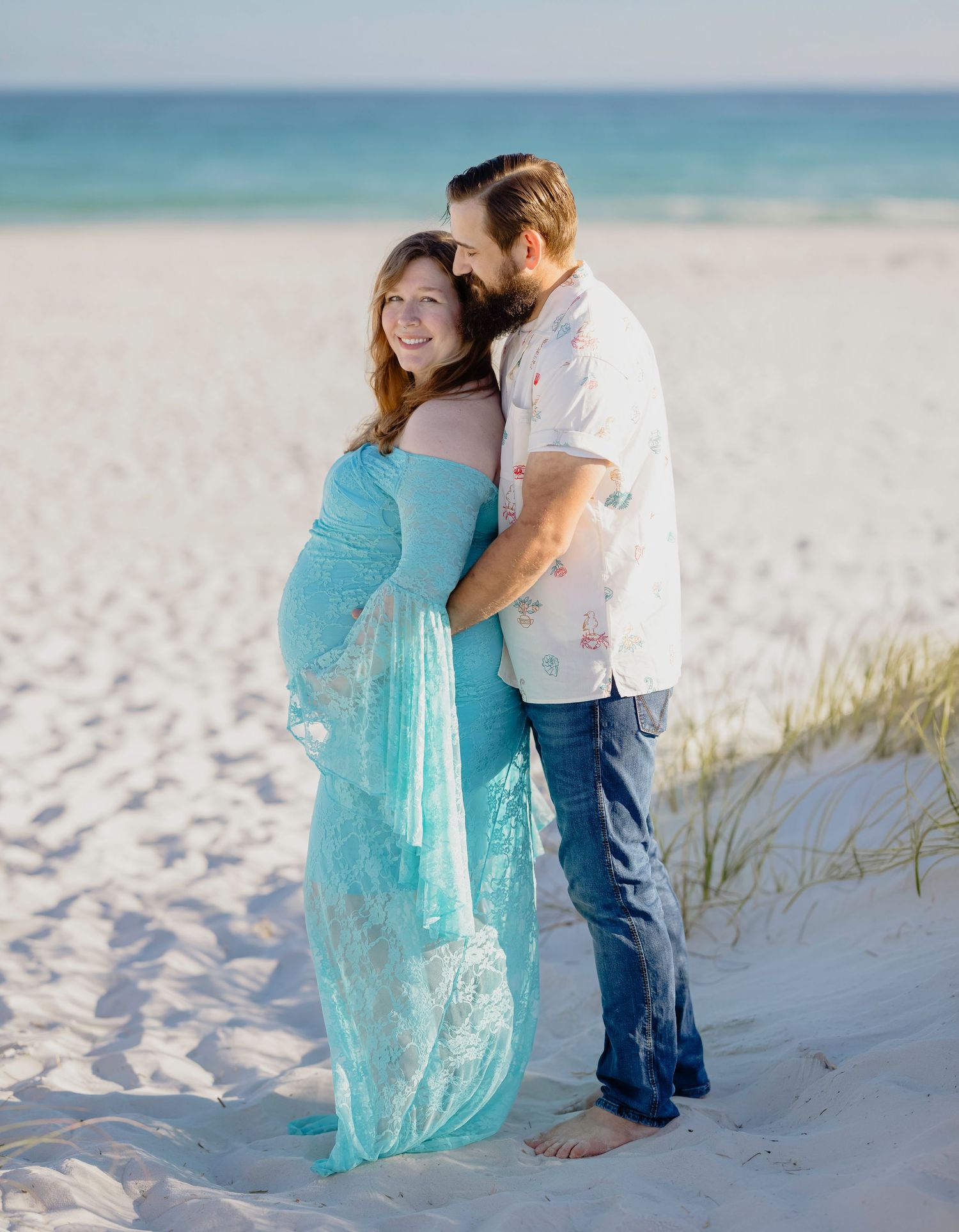 Expecting couple embracing on white sand beach, woman in flowing teal lace maternity gown.