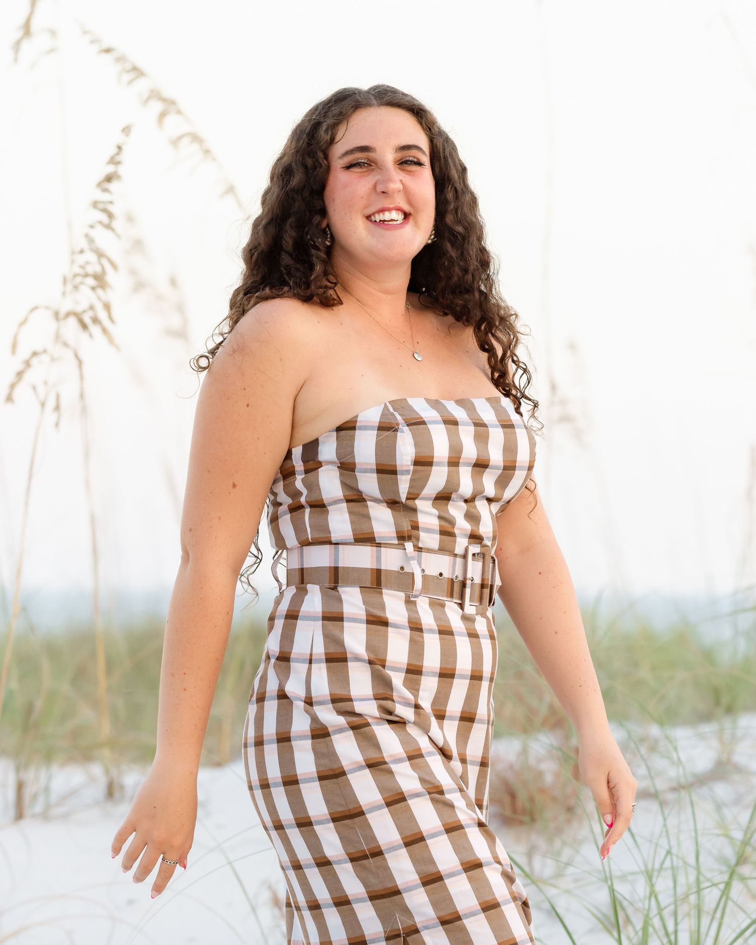Young woman in brown and white checkered strapless dress posing on white sand beach with sea oats.