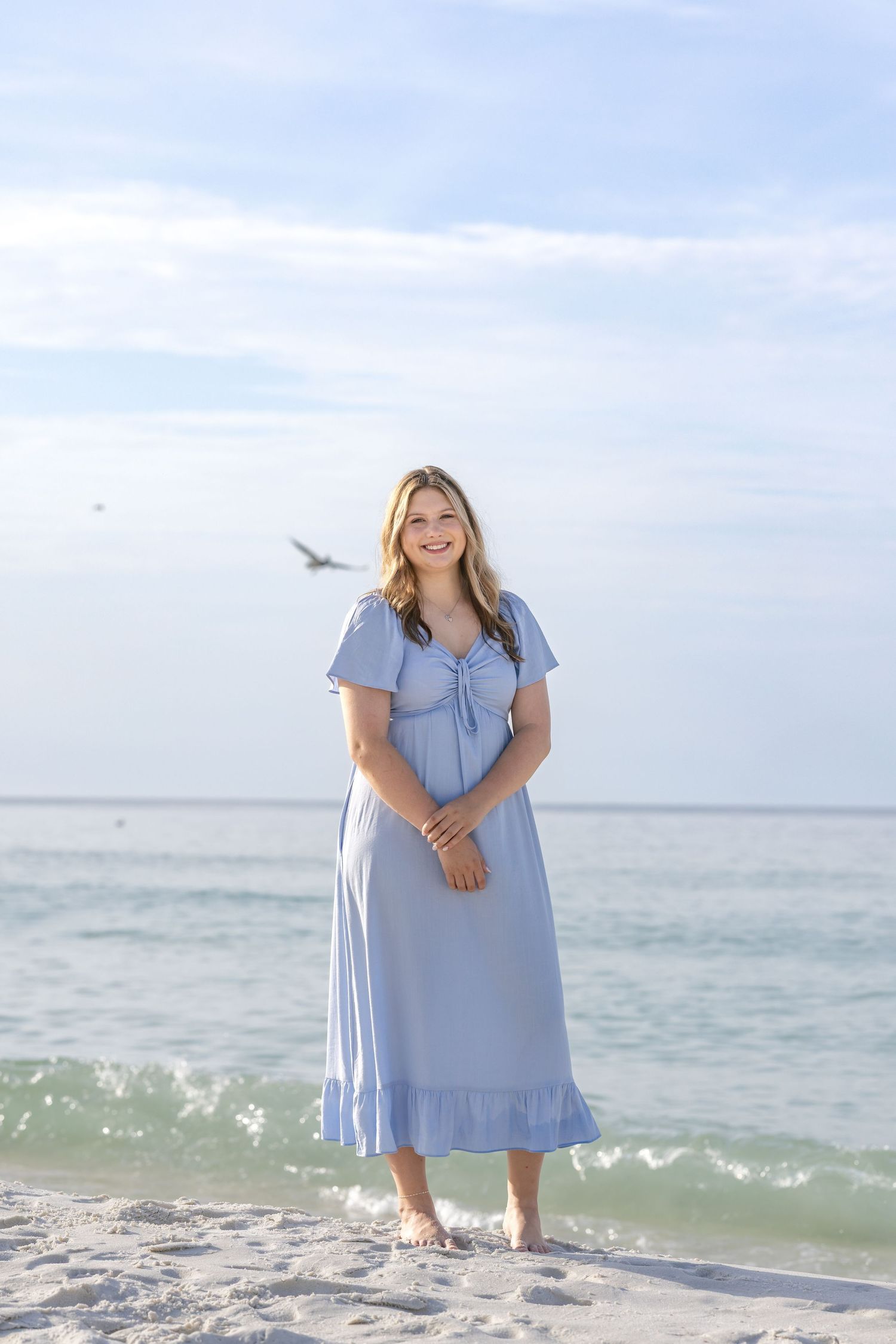 Young woman in a light blue midi dress standing barefoot on a white sand beach with ocean waves behind her.