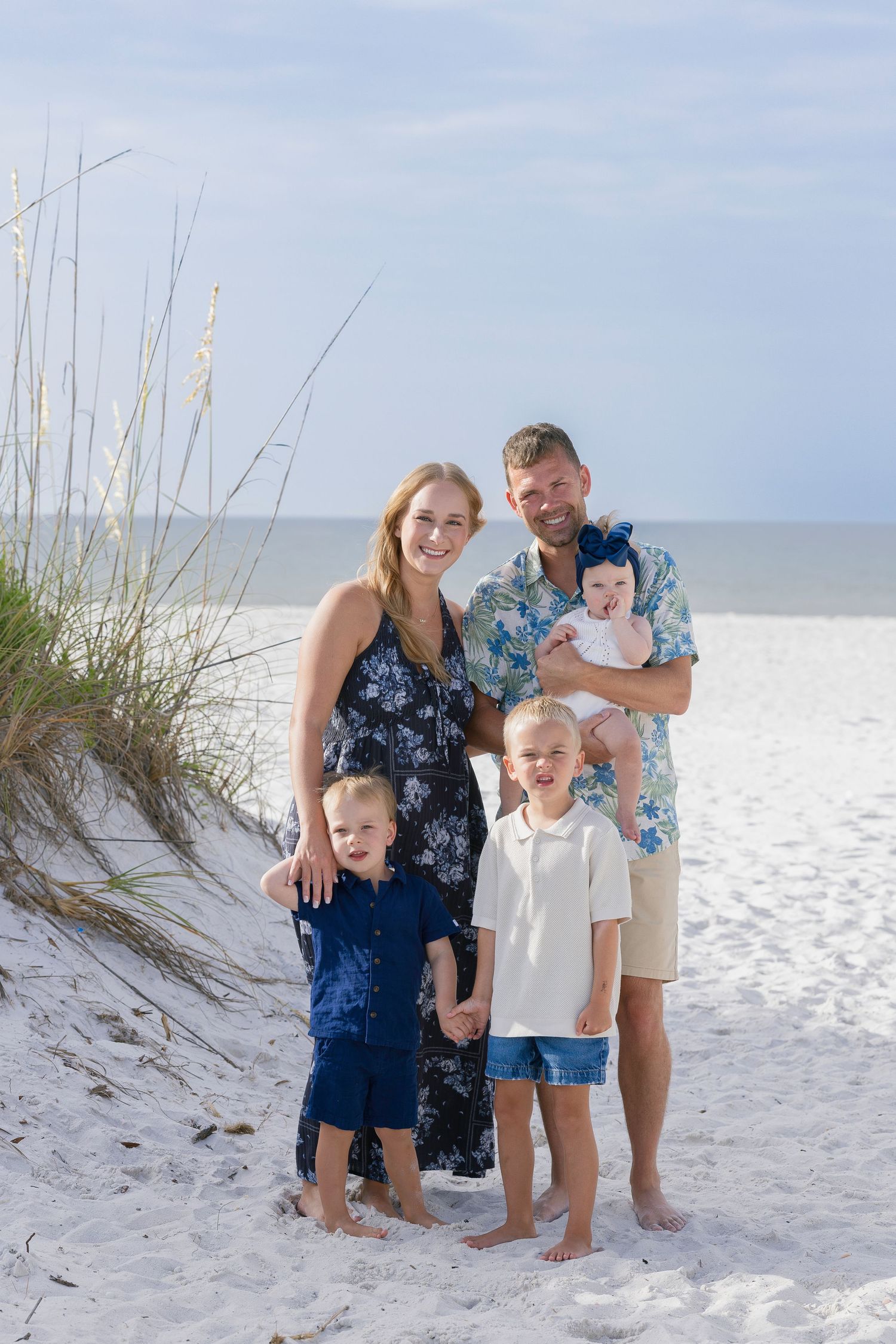 Family of five posing on white sand beach, parents with three children including baby, sea oats in background.