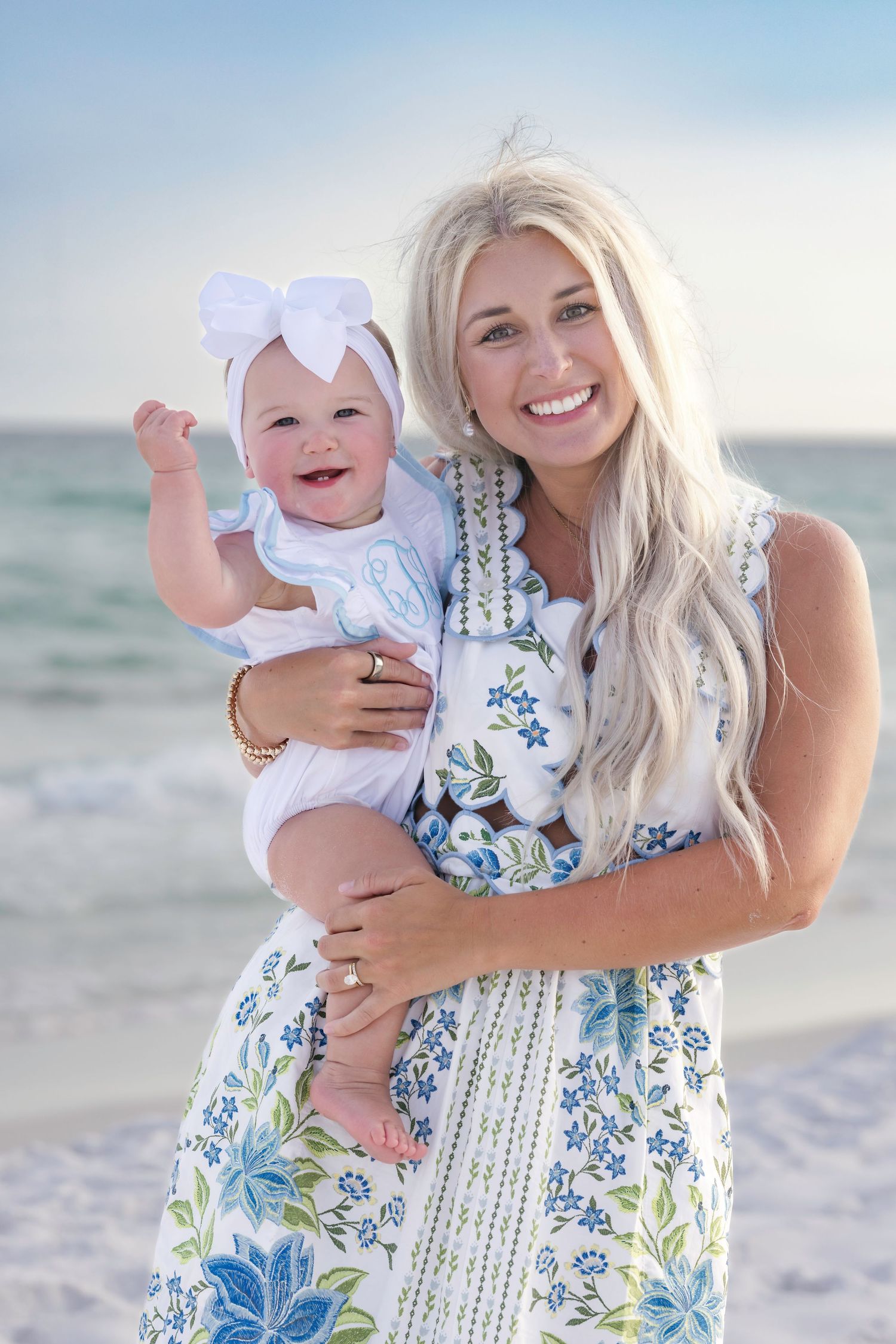 Smiling blonde woman holding a happy baby with a white bow on a sunny beach.
