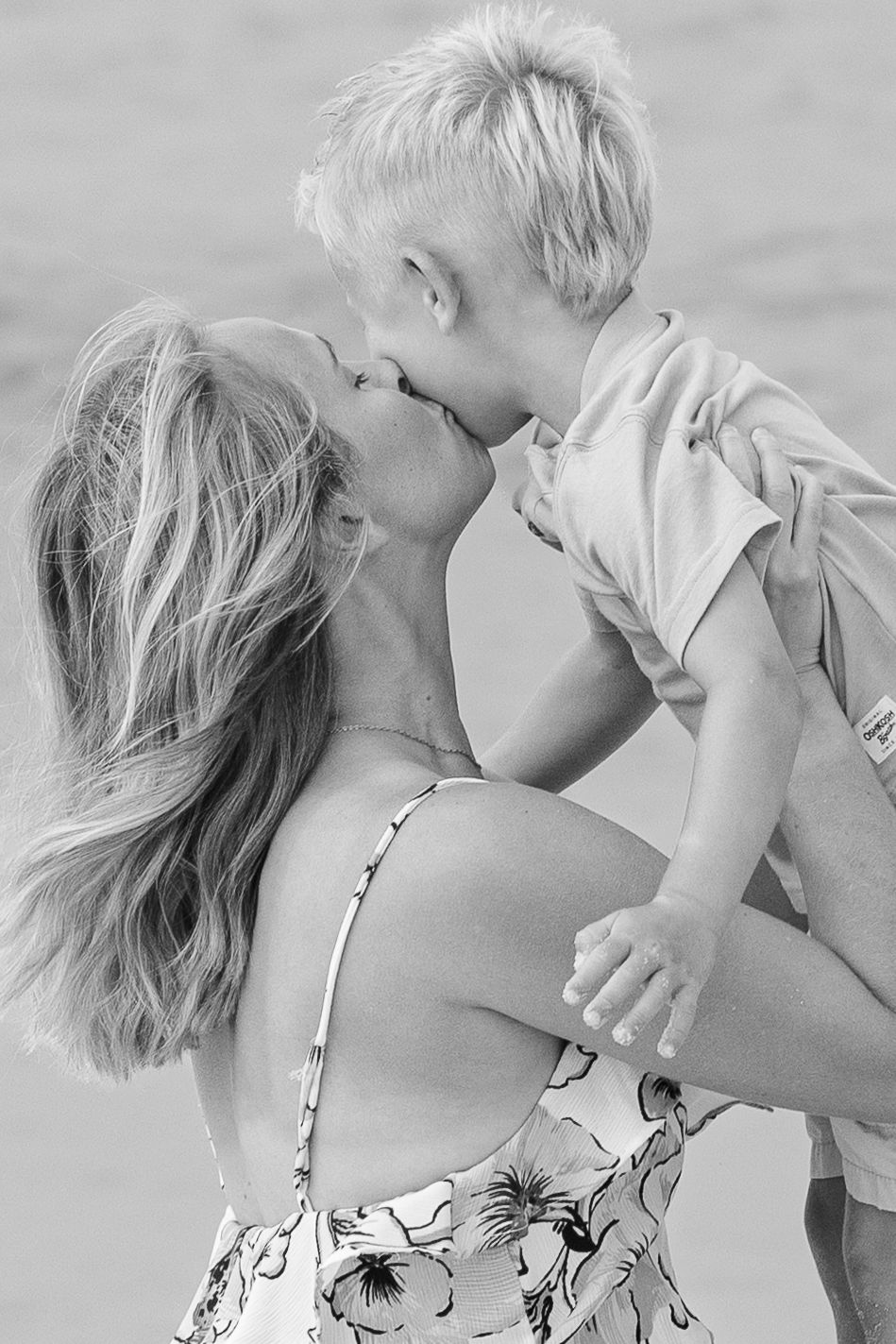 Black and white photo of a mother kissing her young blonde son while holding him at the beach.