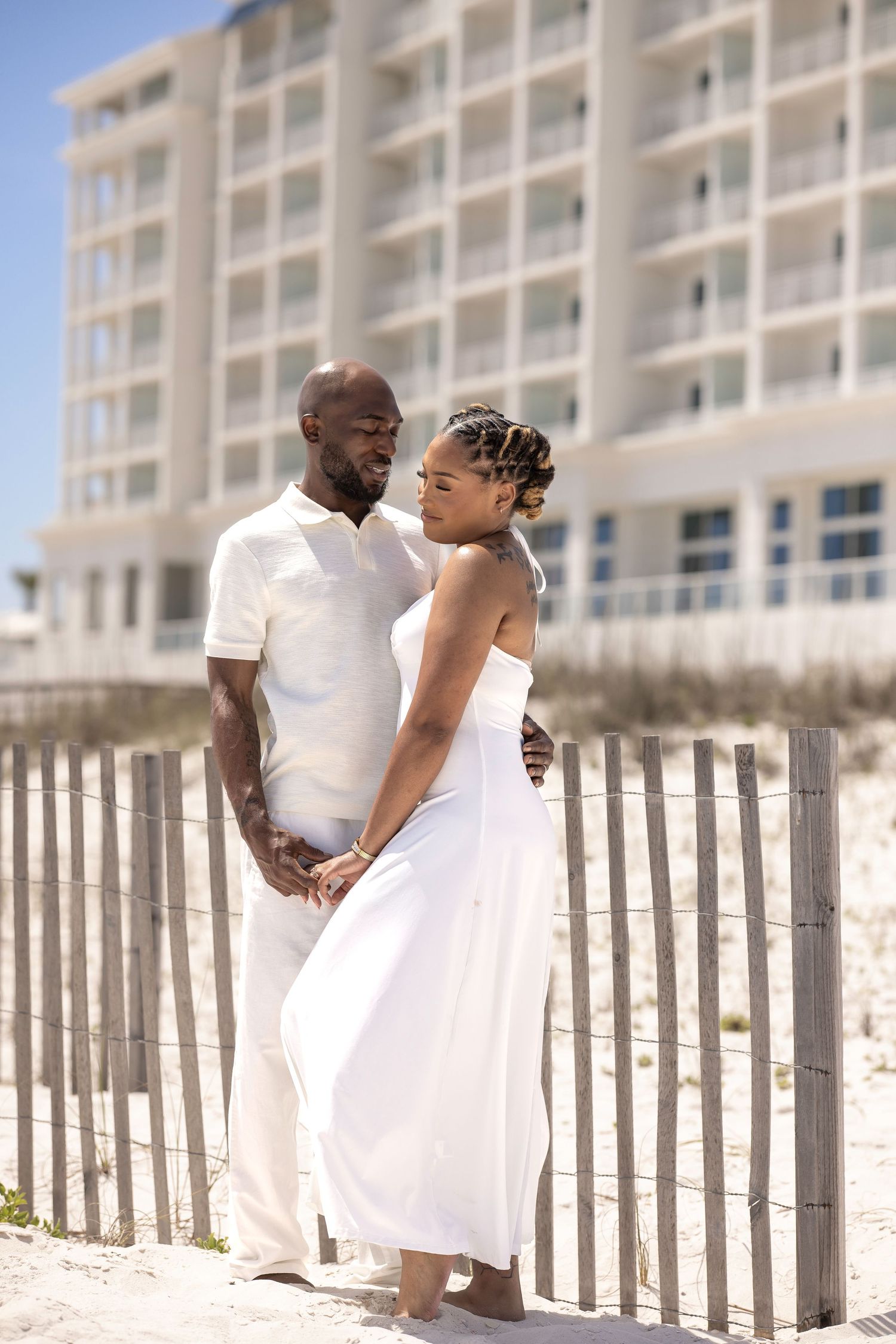 Couple in white outfits holding hands on sandy beach path with wooden fence and hotel building in background.