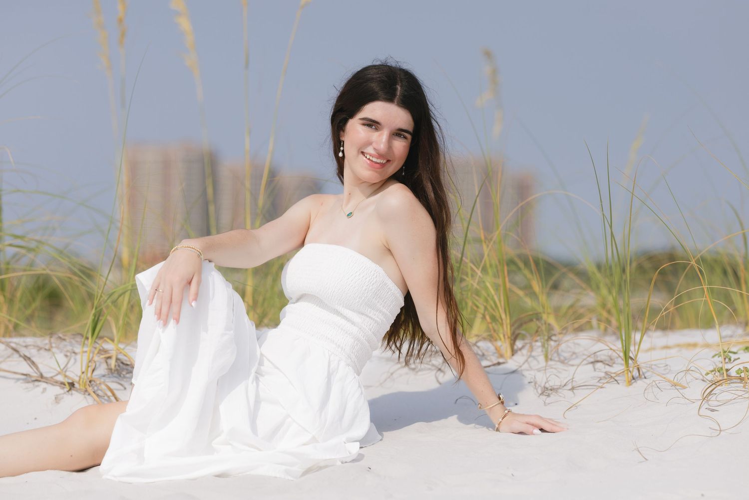 Young woman in white strapless dress sitting on white sand beach dunes with tall grass and buildings in background.