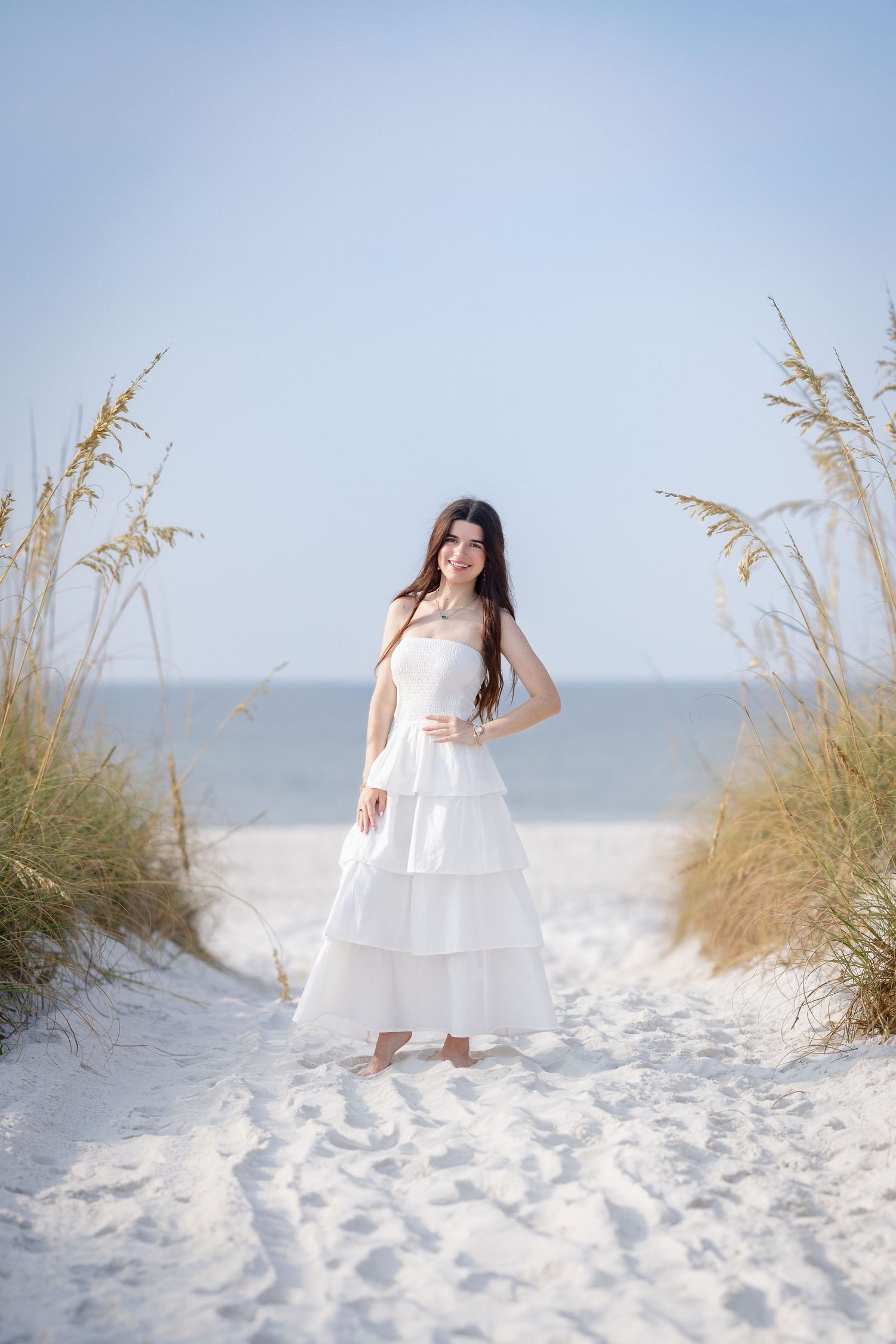 Young woman in white tiered dress standing on sandy beach path flanked by sea oats.