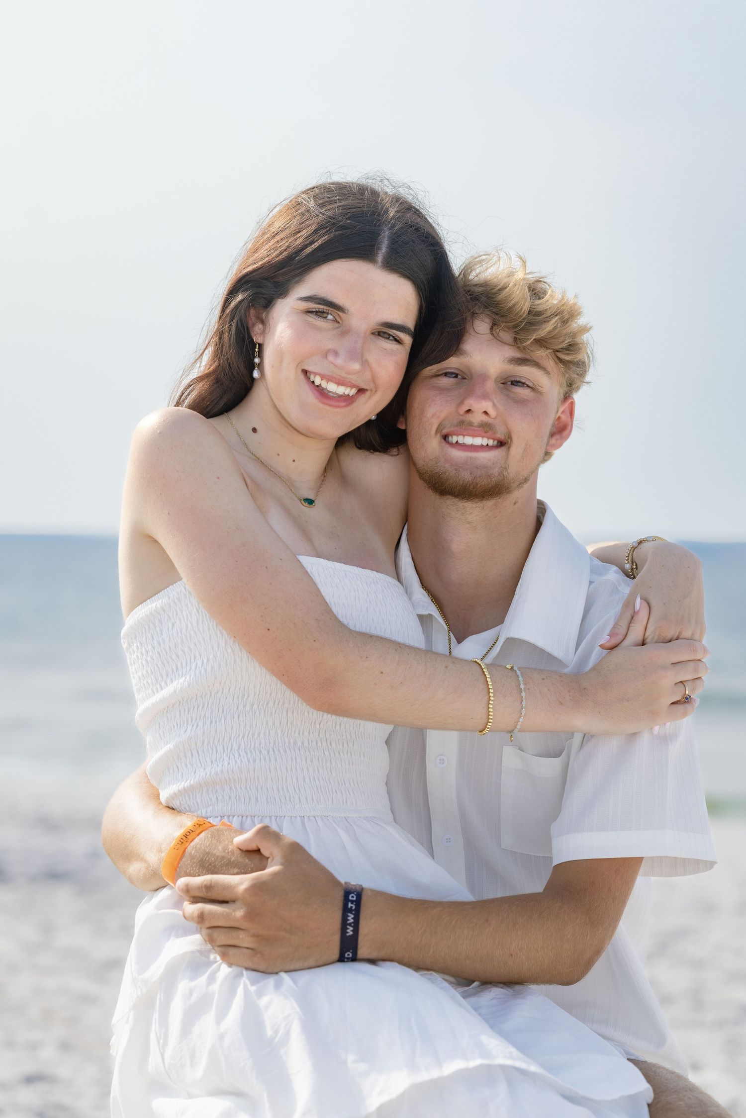 Young couple embracing and smiling on a sunny beach, dressed in white outfits with the ocean in the background.