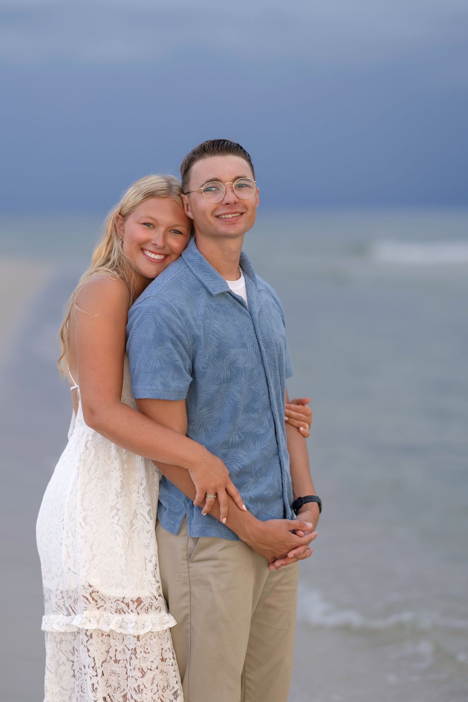 Happy couple embracing on a beach, woman in white lace dress and man in blue shirt, with ocean waves in the background.