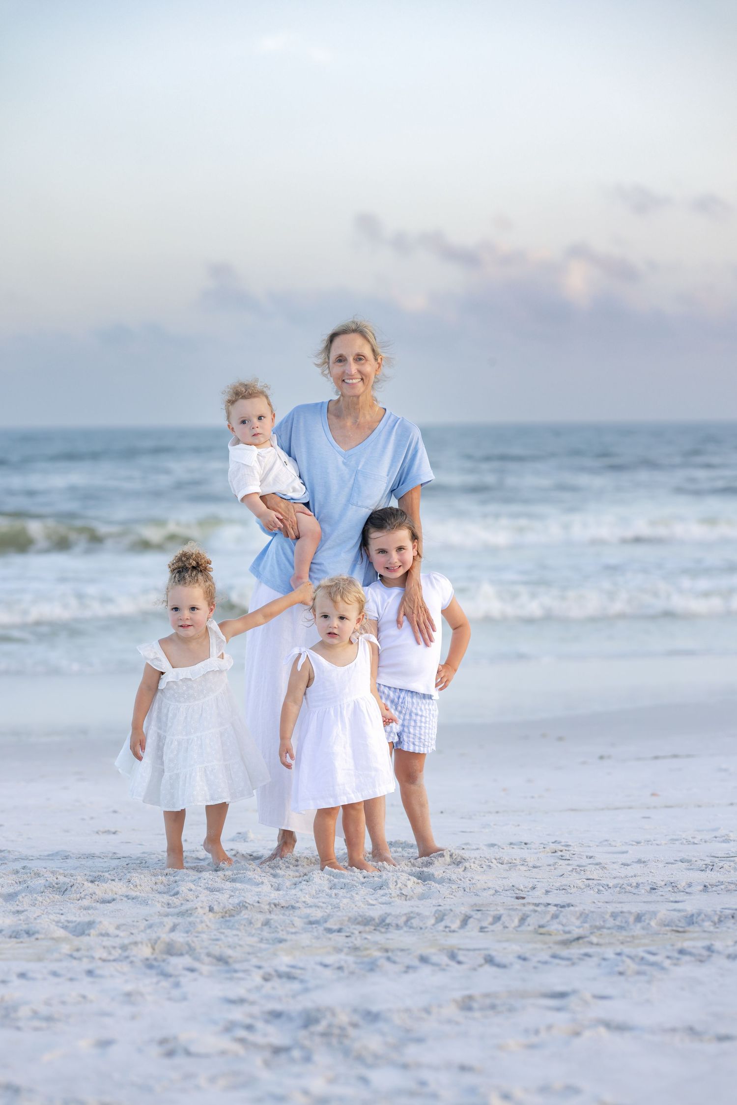 Woman holding baby poses with four young children in white outfits on a sandy beach at sunset.