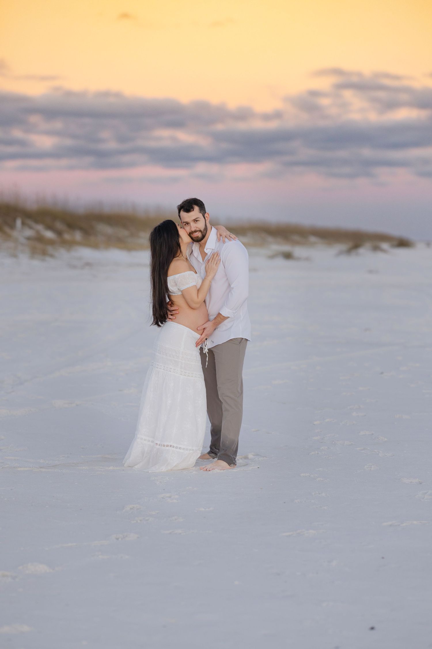 A couple shares a romantic moment on a pristine white sand beach at sunset with soft orange and purple sky.