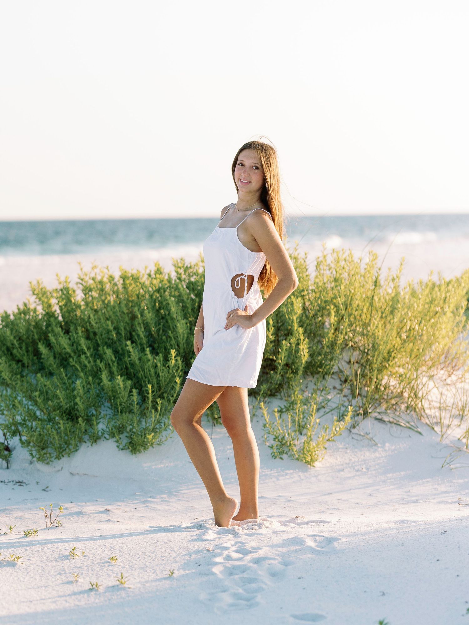 Woman in white outfit poses on sandy beach with sea grass and ocean in background.