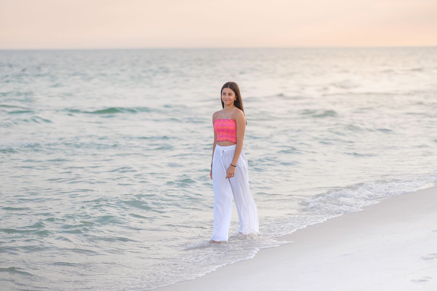 Person in white dress walking along a serene beach at sunset with ocean waves in the background.