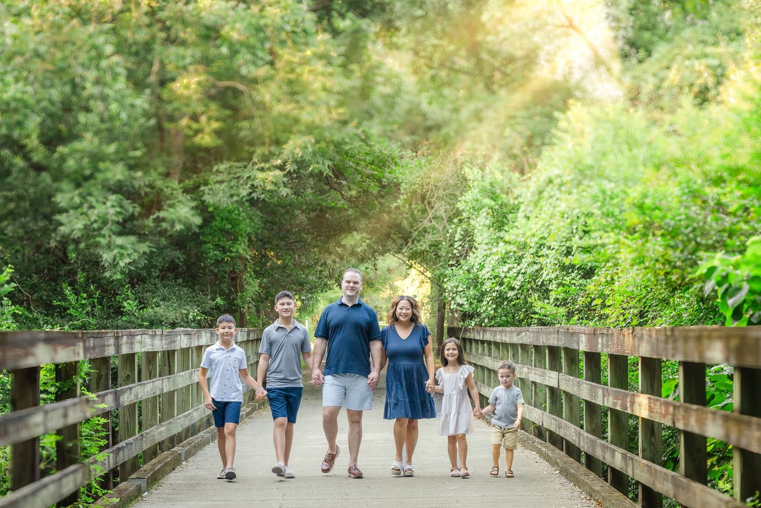 Group walks together holding hands along a wooden boardwalk surrounded by lush green foliage on a sunny afternoon.