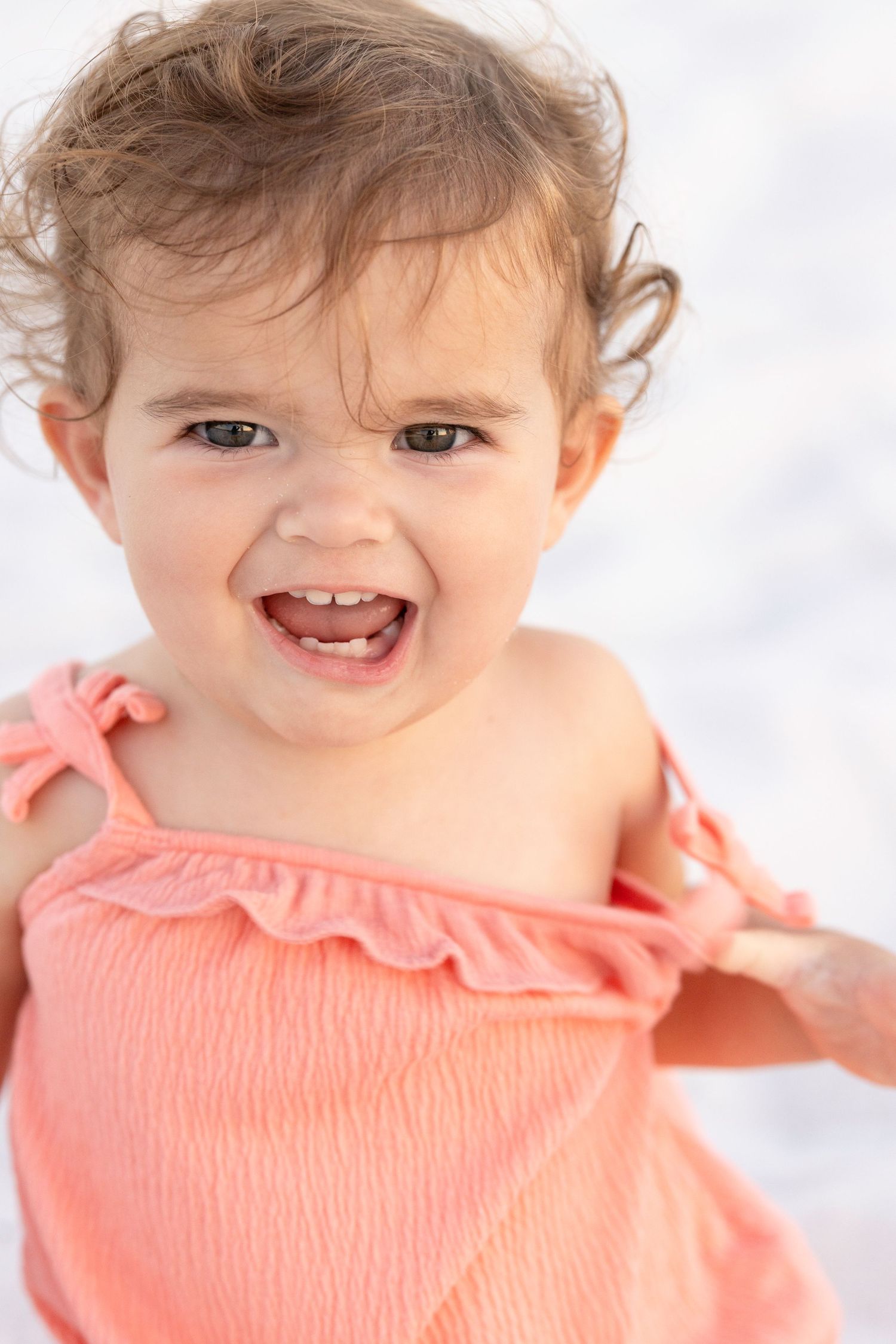 A joyful toddler wearing a coral-colored dress laughs while posing against a white background.