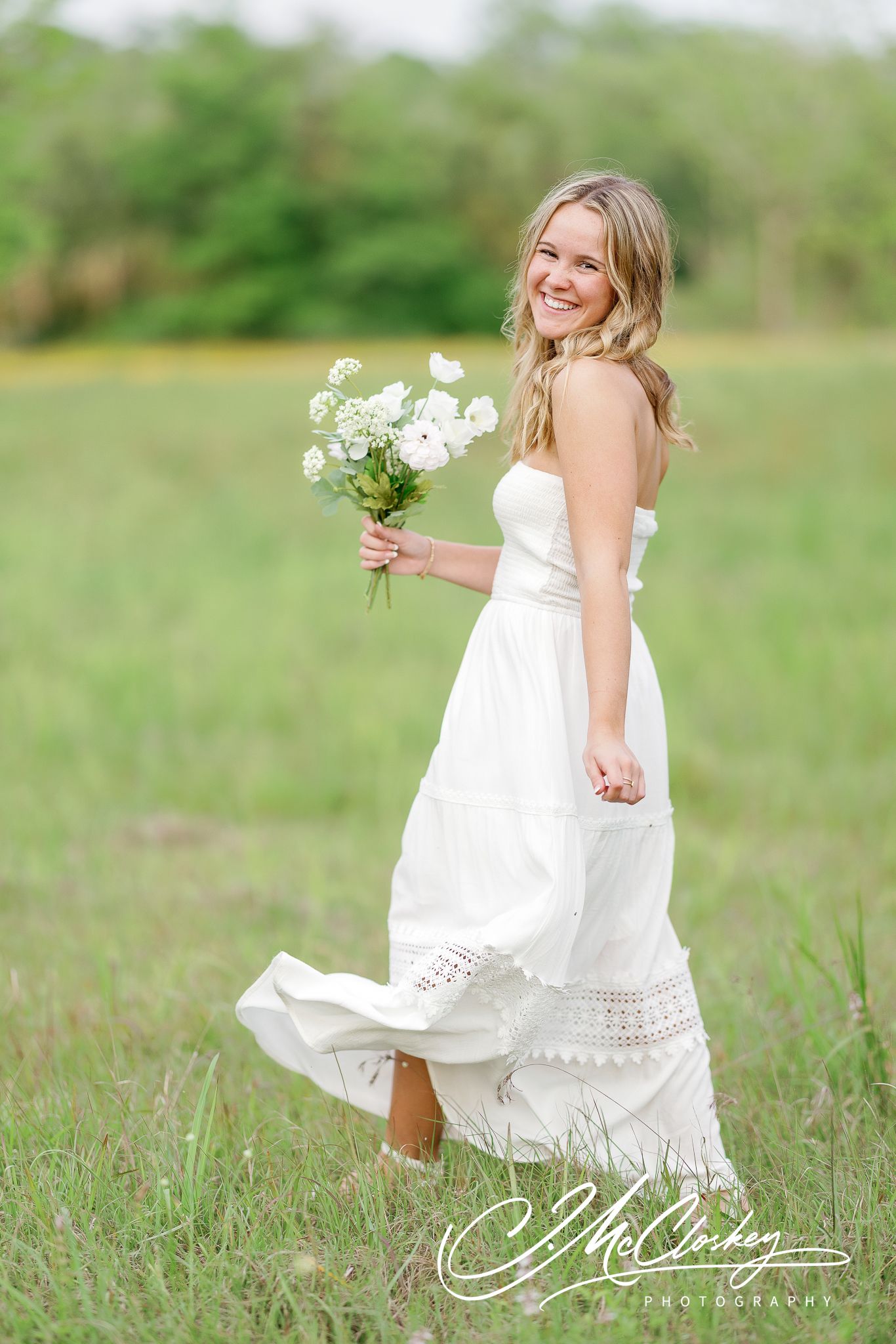 A woman in a flowing white dress twirls and dances in a green spring meadow while holding wildflowers.