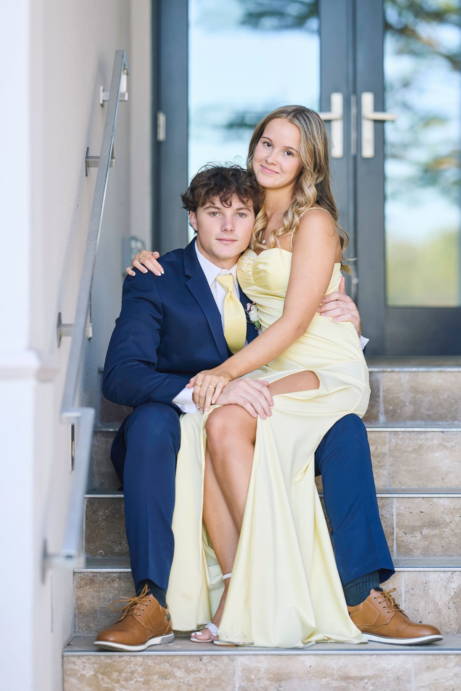 A couple sits together on outdoor stone steps, with the person wearing a navy suit and brown shoes positioned to one side.