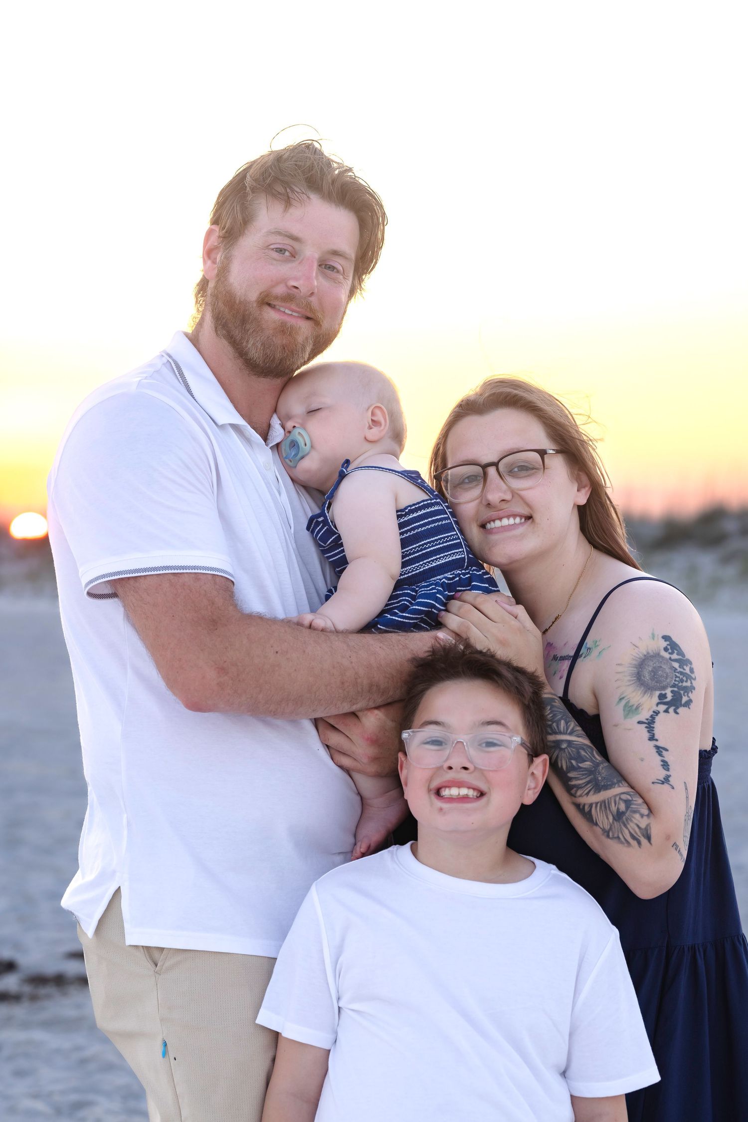 Smiling family of four at beach sunset, bearded dad holding baby, mom with tattoos, young boy in white tee.