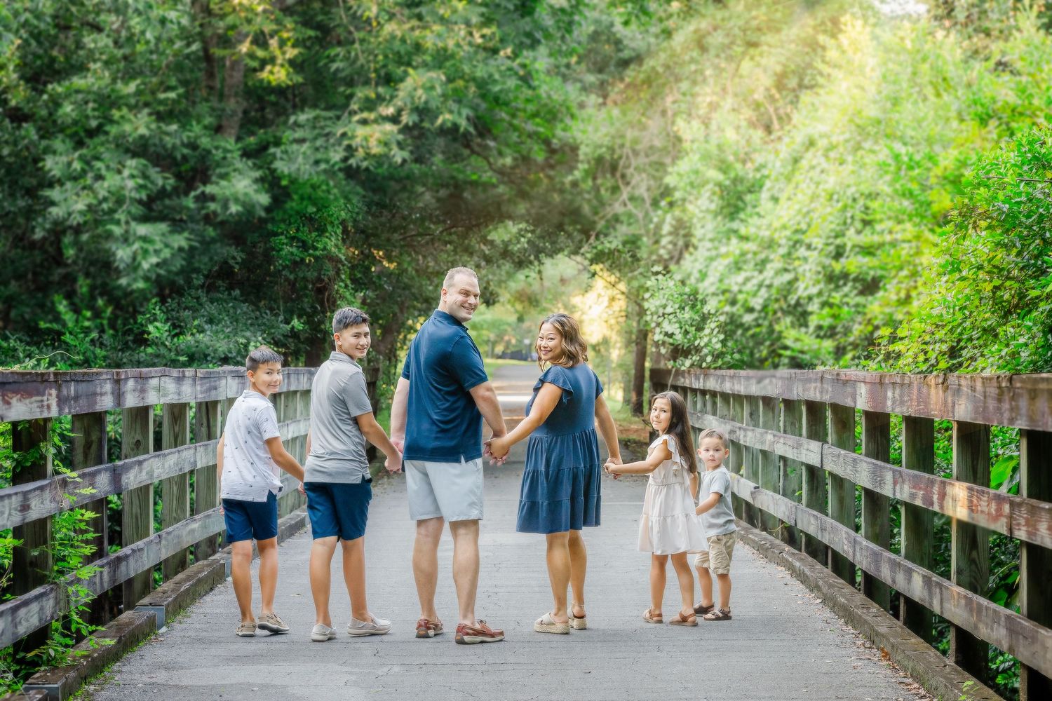 Family of six holding hands on a wooden bridge surrounded by lush green trees on a sunny day.