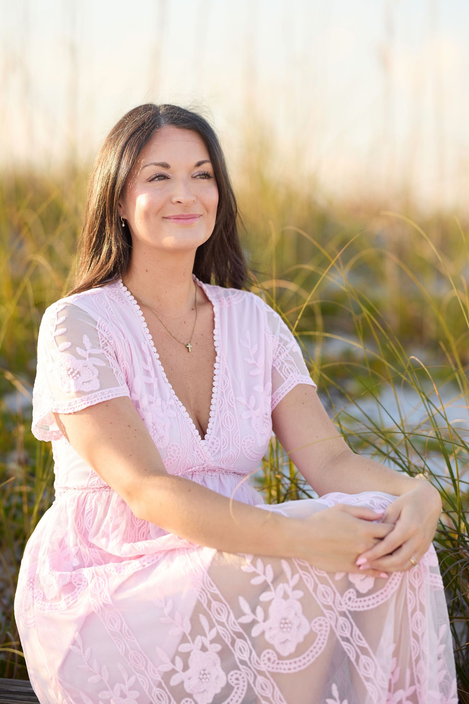 Woman in pink floral dress sitting outdoors near tall grass, smiling warmly in natural golden light.