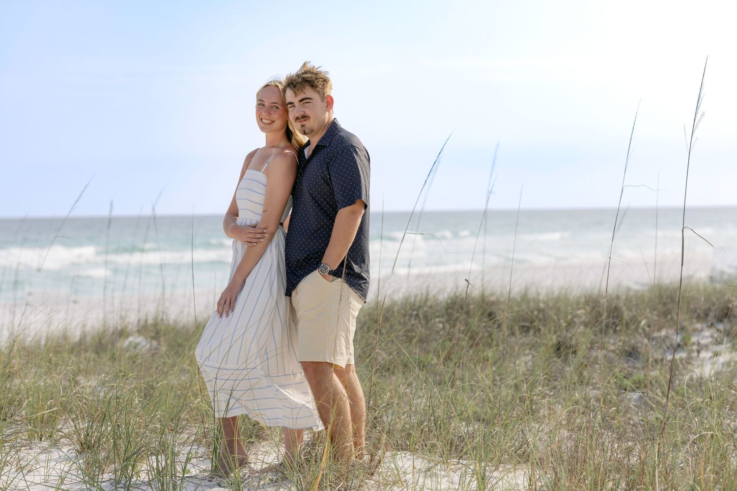 Couple embracing on a sunny beach with ocean waves and sea grass in the background.