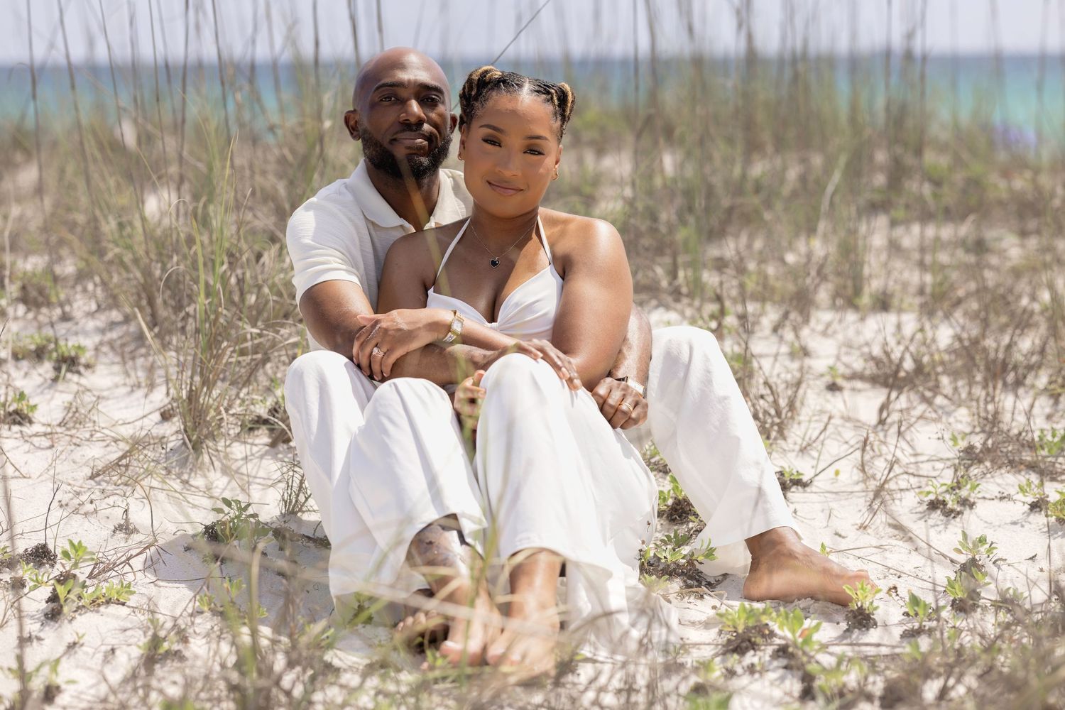 Couple in white outfits sitting together on sandy beach with ocean in background.
